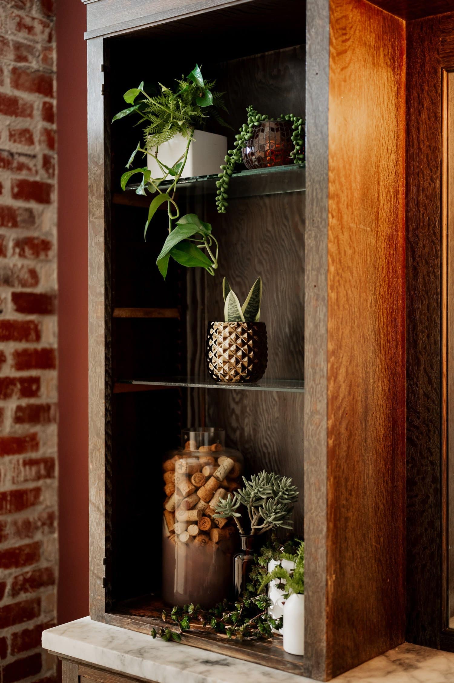 A wooden cabinet with various lush plants, including a potted succulent, standing against a brick wall.