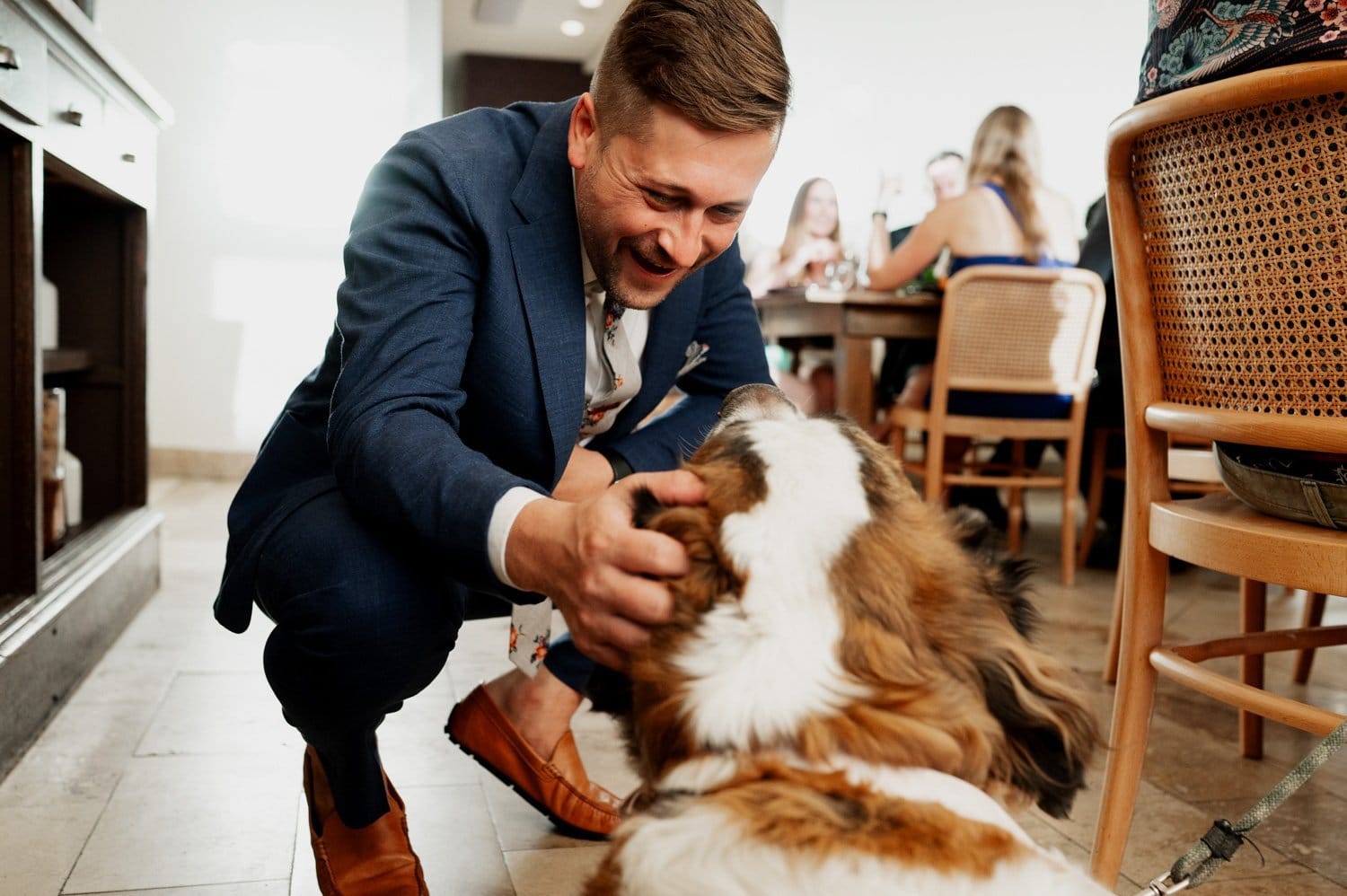 Man in a blue suit happily petting a large dog indoors, with guests seated at a dining table in the background during a wedding at Opal 28.