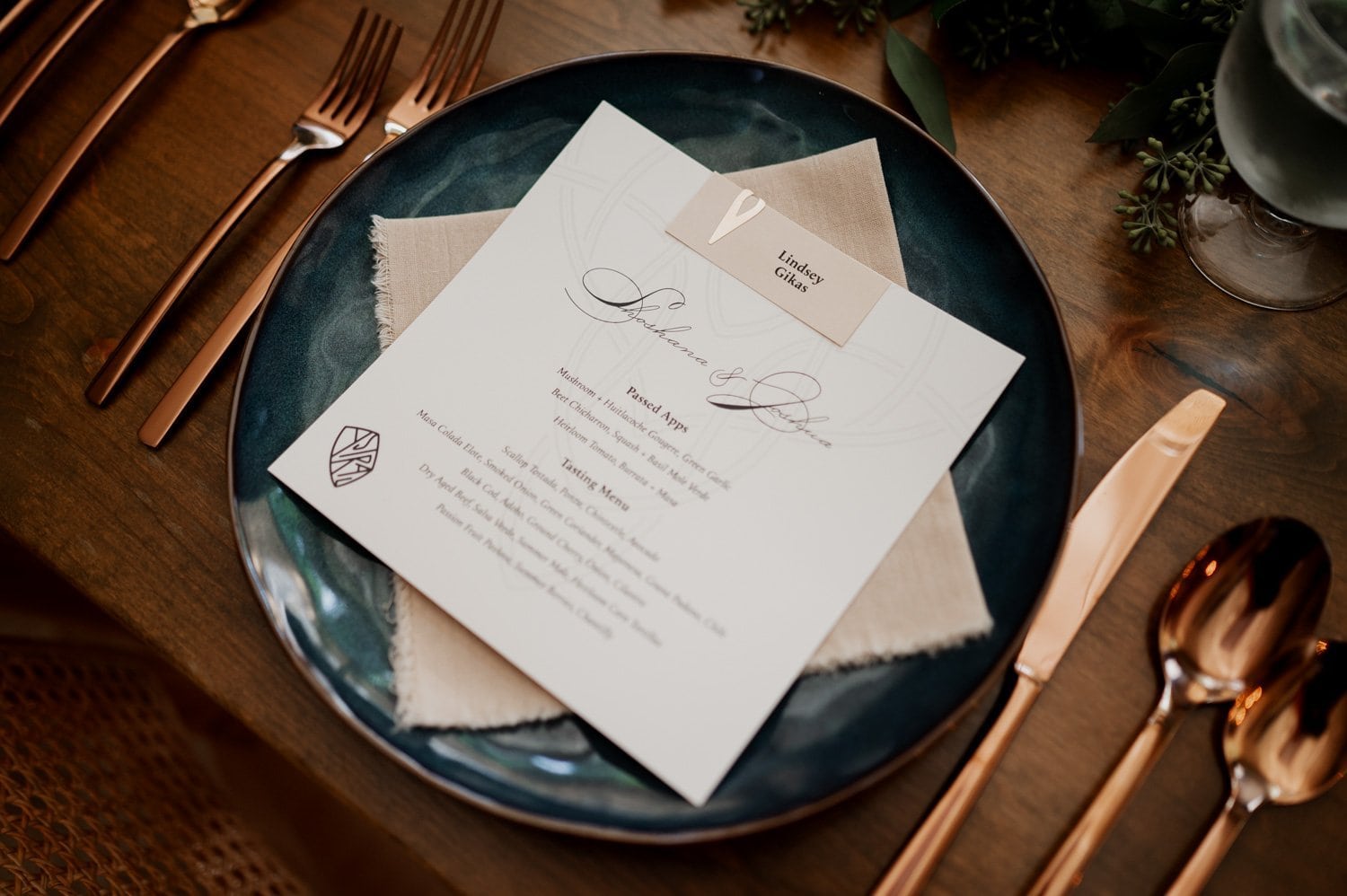 Elegant wedding table setting at Opal 28 with a menu placed on a blue plate, surrounded by copper cutlery and lush greenery accents.