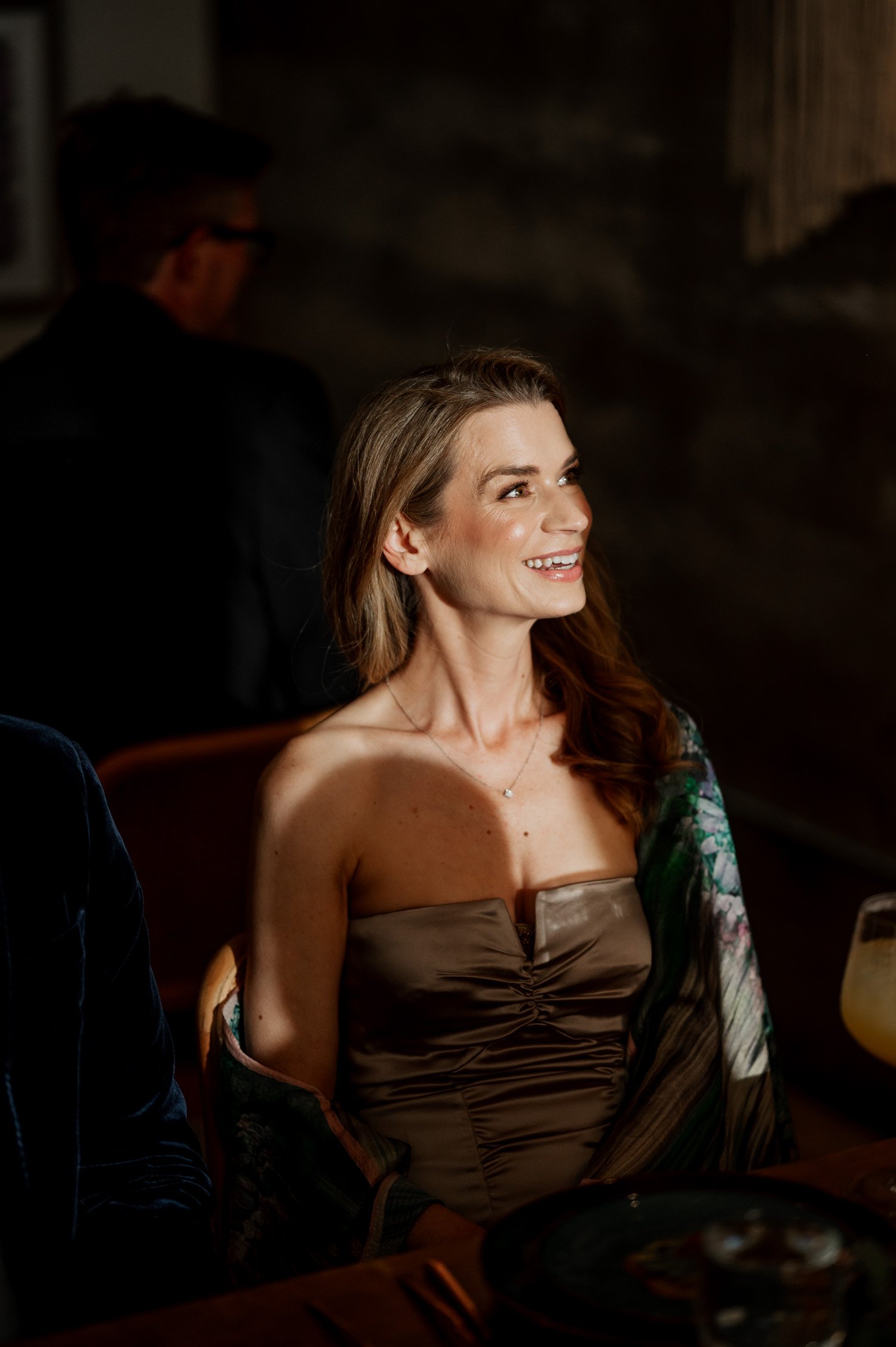 A woman smiles while sitting at a dimly lit table in a restaurant, holding a drink in front of her. She appears to be lost in thought, possibly reflecting on fond memories.