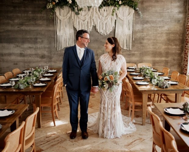 A couple in wedding attire stands hand in hand, surrounded by wooden tables and macramé hangings in a decorated room at Opal 28.