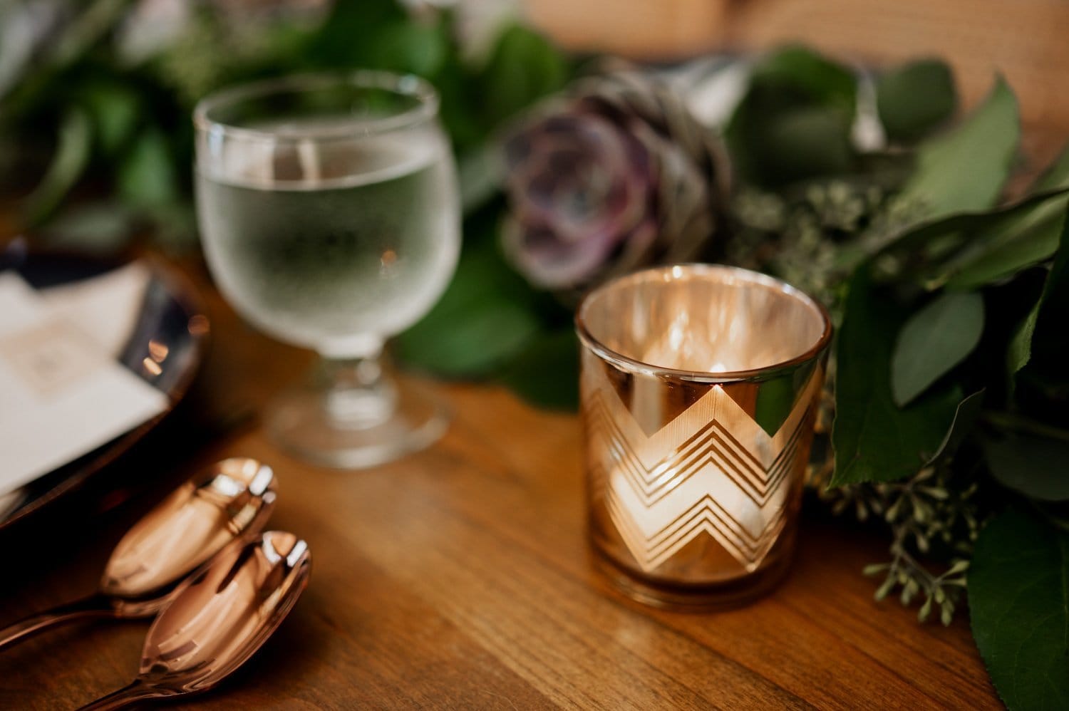 A decorative candle, a glass of water, and golden spoons are placed on a wooden table, with greenery around them, creating a picturesque setting for a wedding at Opal 28.