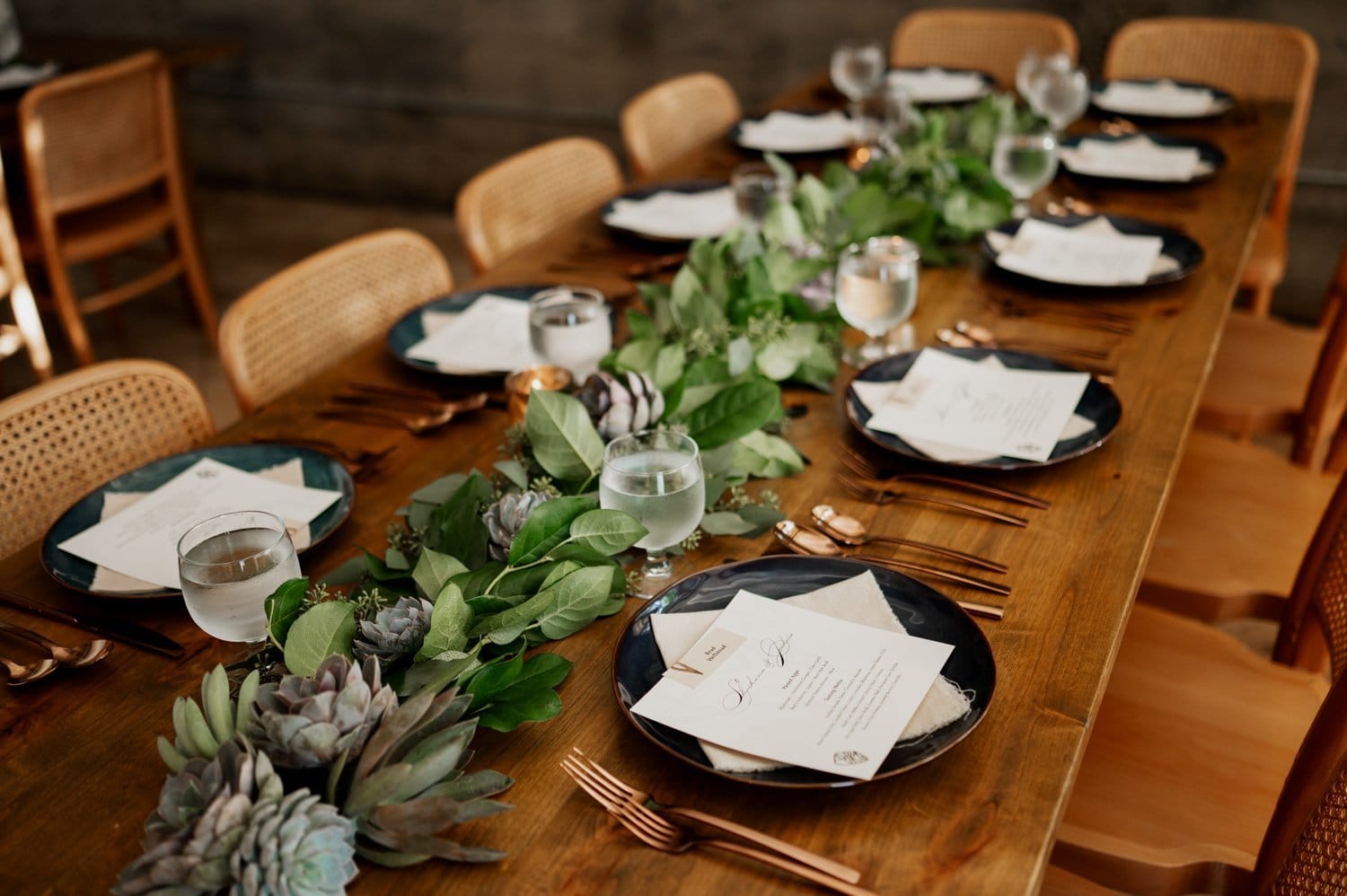 A beautifully arranged dining table featuring neatly set plates, menus, and glasses of water, complemented by a lush green foliage centerpiece. The ambiance is warm and inviting, ideal for a wedding at Opal 28.