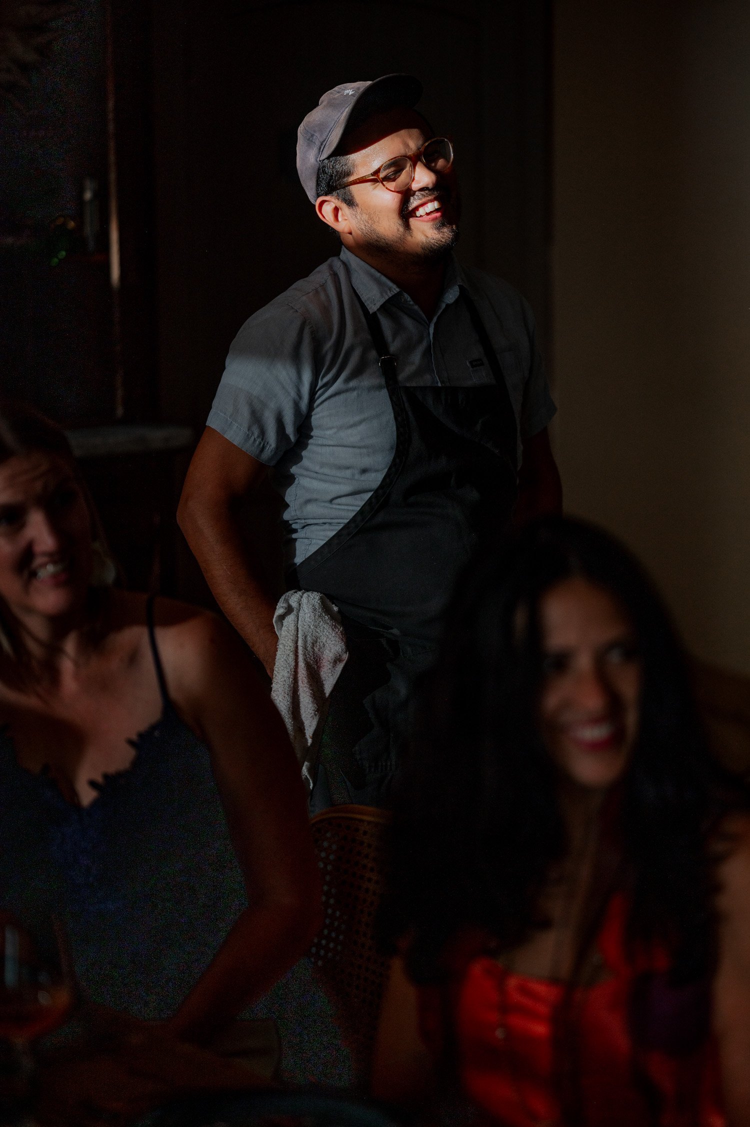 In the softly illuminated room, a man wearing a cap and apron smiles warmly at two seated women, creating a joyful atmosphere.