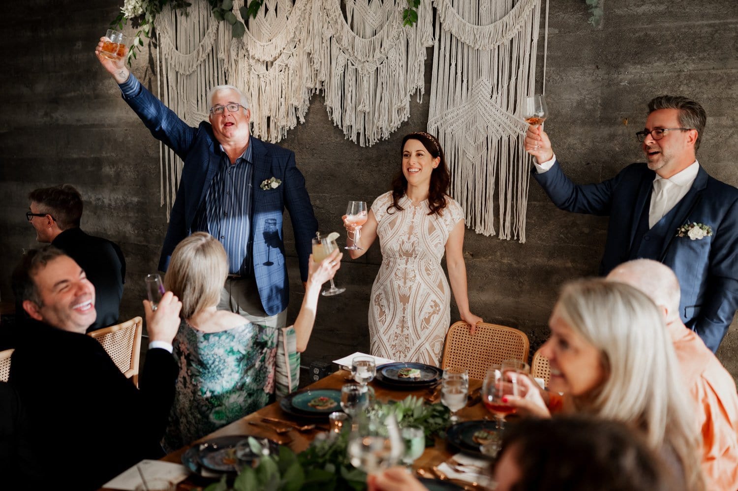 A joyful group raises glasses in a toast at a wedding reception, smiling near a macramé backdrop and celebrating the occasion at Opal 28.