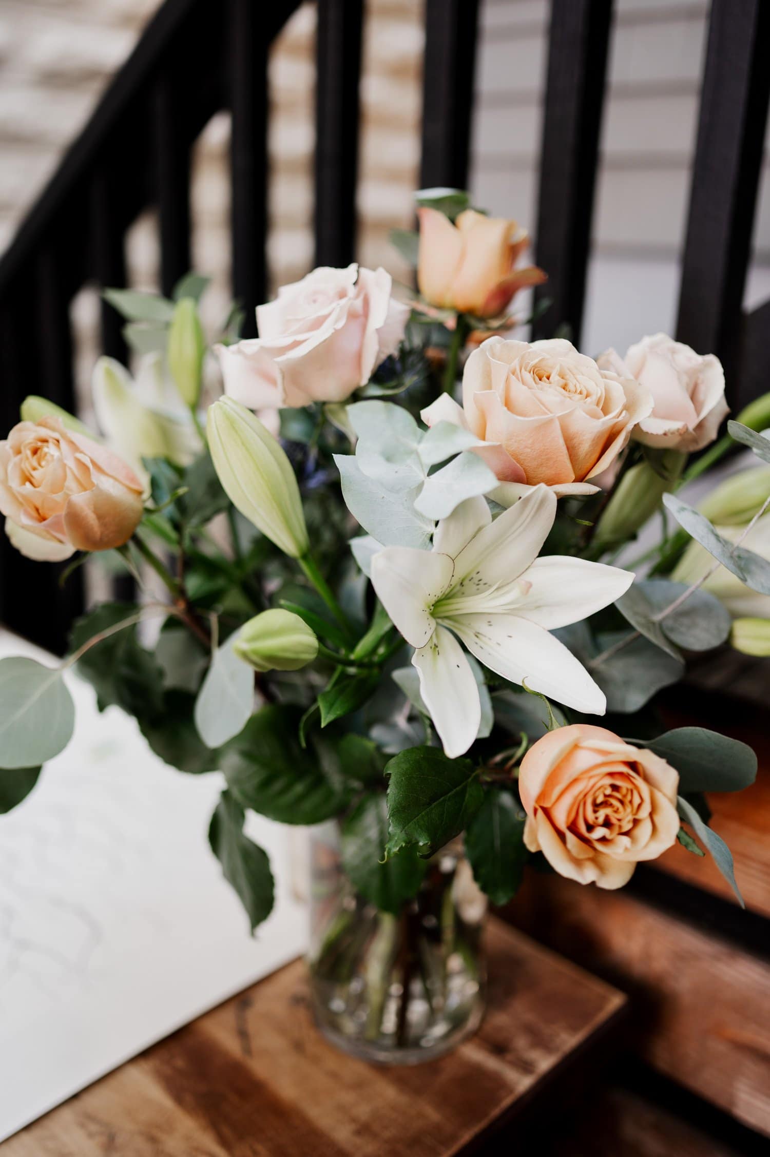 A bouquet of pale pink roses, white lilies, and green leaves arranged in a glass vase on a wooden surface.