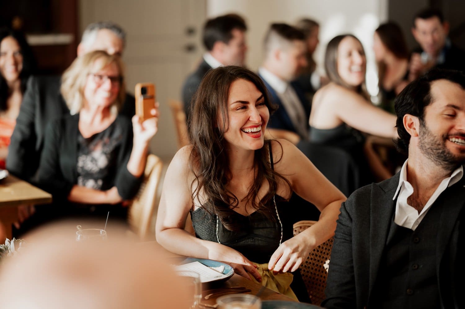 At a wedding at Opal 28, a woman bursts into laughter, bringing cheer to the formal event as guests seated around her join in the joy.