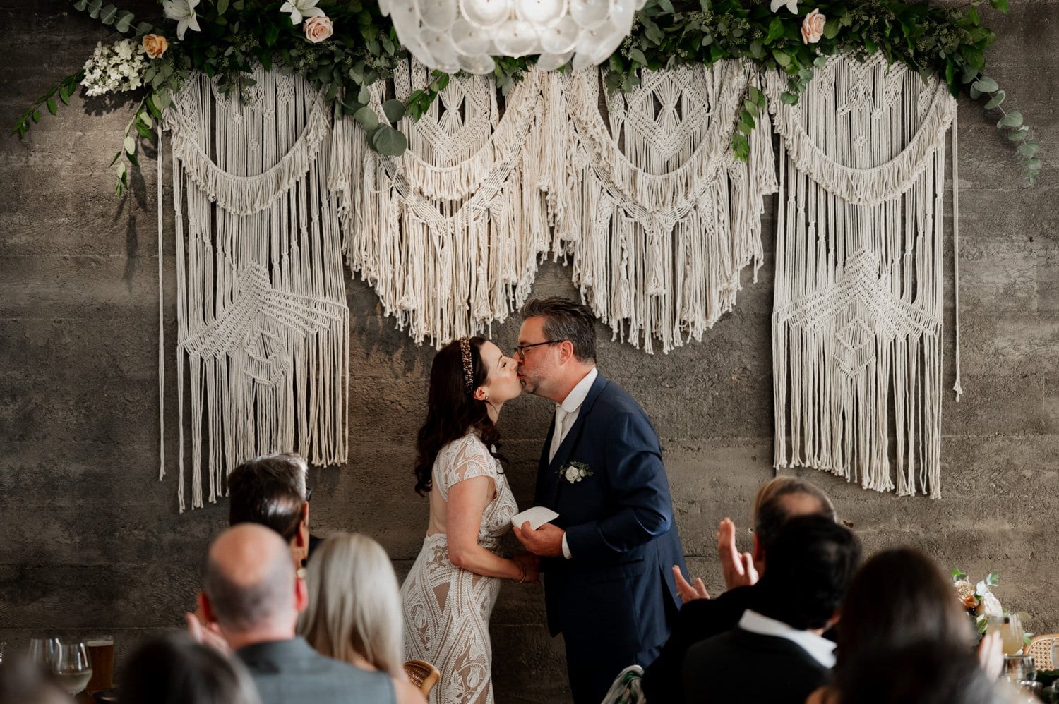 The couple, dressed in elegant wedding attire, share a kiss beneath macramé decor at their wedding at Opal 28, while guests watch and clap joyfully.