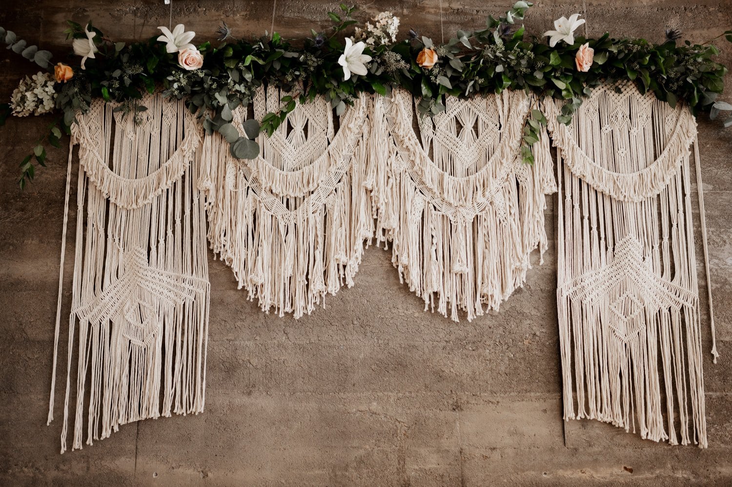 Four macramé wall hangings with floral garlands decorate the stone wall, creating a charming backdrop perfect for a wedding at Opal 28.