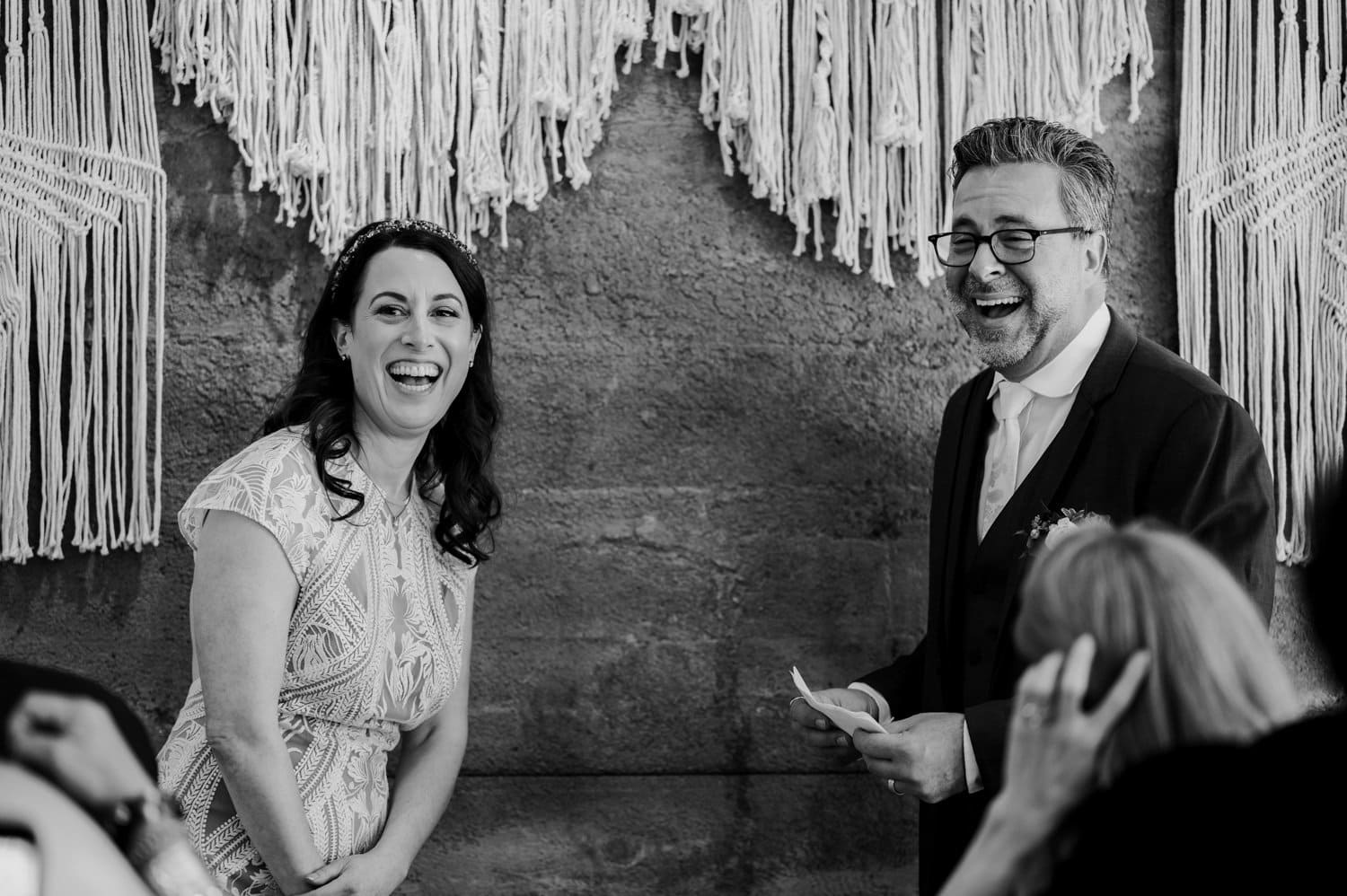 Bride and groom laughing together at a wedding ceremony with a macramé backdrop at Opal 28.