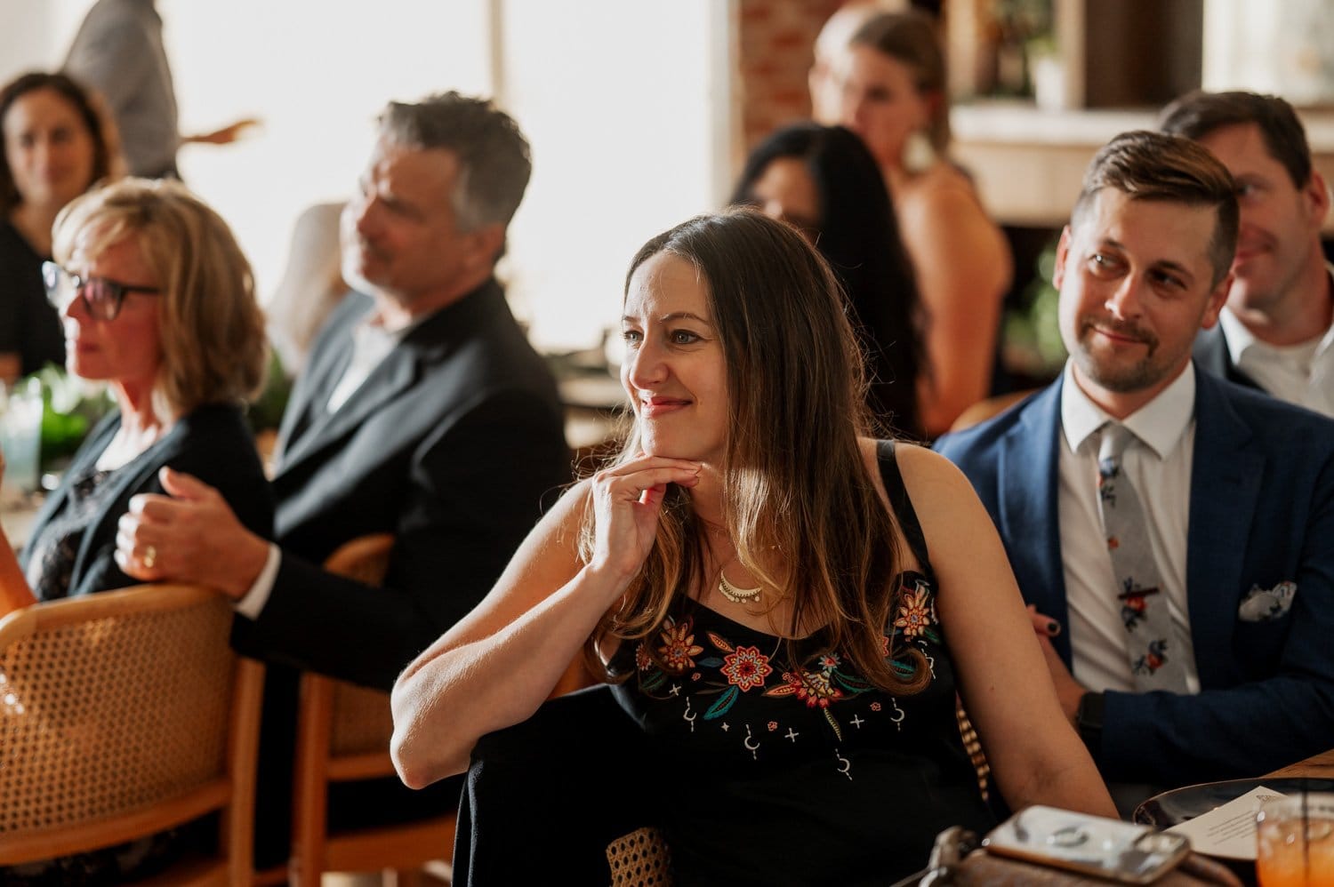 A group of people sitting and smiling warmly at a wedding at Opal 28, with a woman with long hair in the foreground as the focal point.