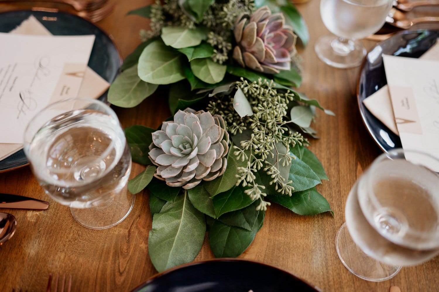 A wedding table at Opal 28 set with green and purple succulents, leafy greens, glasses of water, and elegant place cards on a wooden table.
