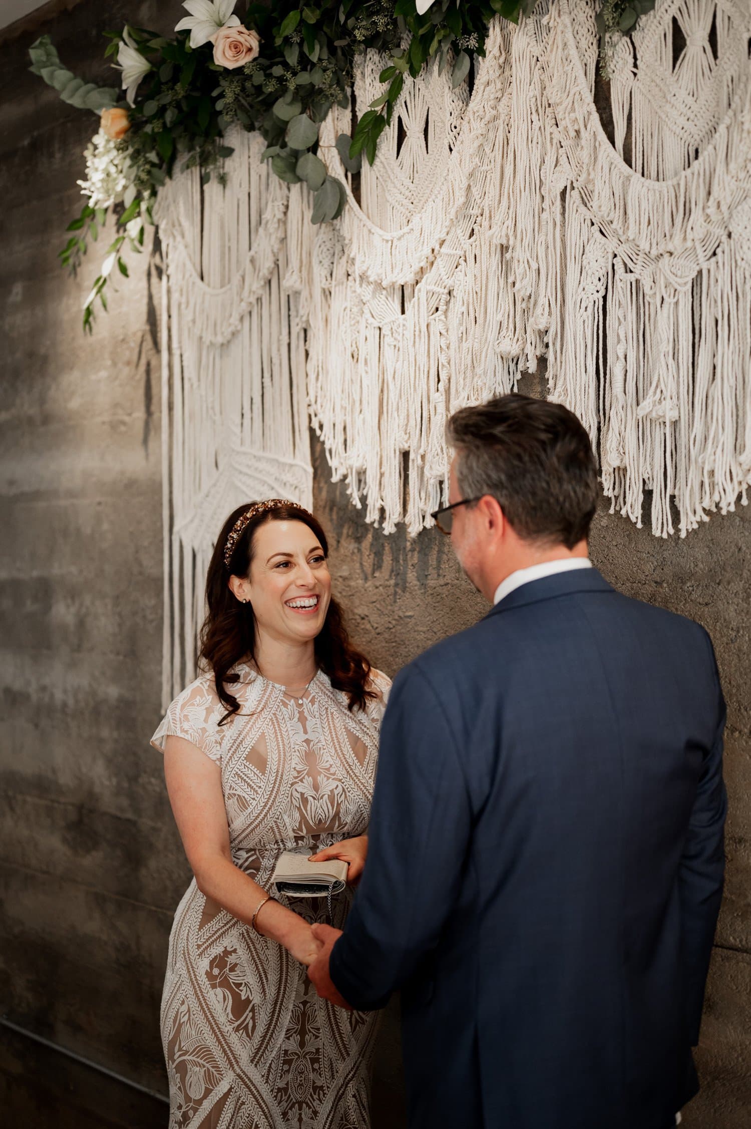 The bride and groom exchange vows under a decorative macramé backdrop with greenery and flowers at their wedding at Opal 28.