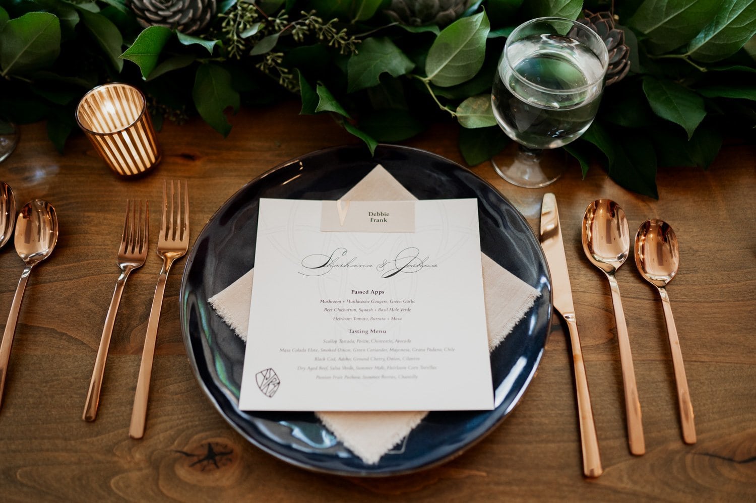 A beautifully arranged table setting for a wedding at Opal 28, showcasing a menu placed on a black plate, accompanied by gold cutlery, a water glass, and a vibrant greenery centerpiece.