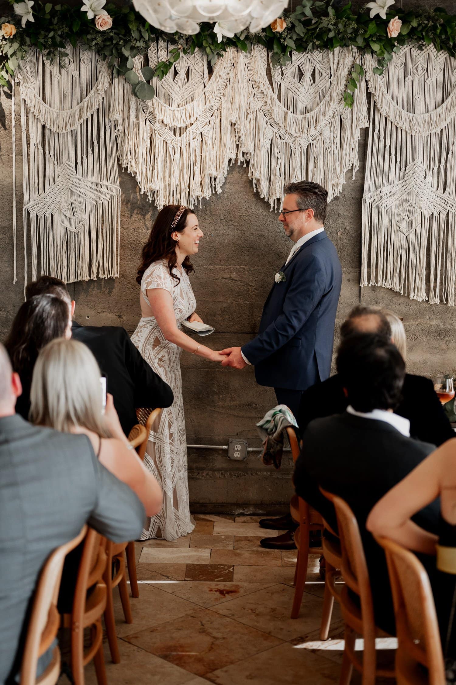 A couple holds hands in front of guests at a bohemian-style wedding, featuring macramé decor and abundant greenery at Opal 28.