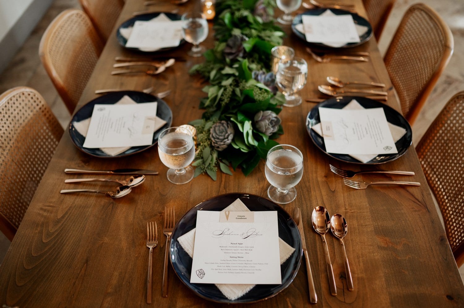 An elegant table setting for a wedding at Opal 28, showcasing menus and dark plates alongside a lush greenery centerpiece. Copper utensils gleam on the wooden table, contributing to the sophisticated and intimate atmosphere.