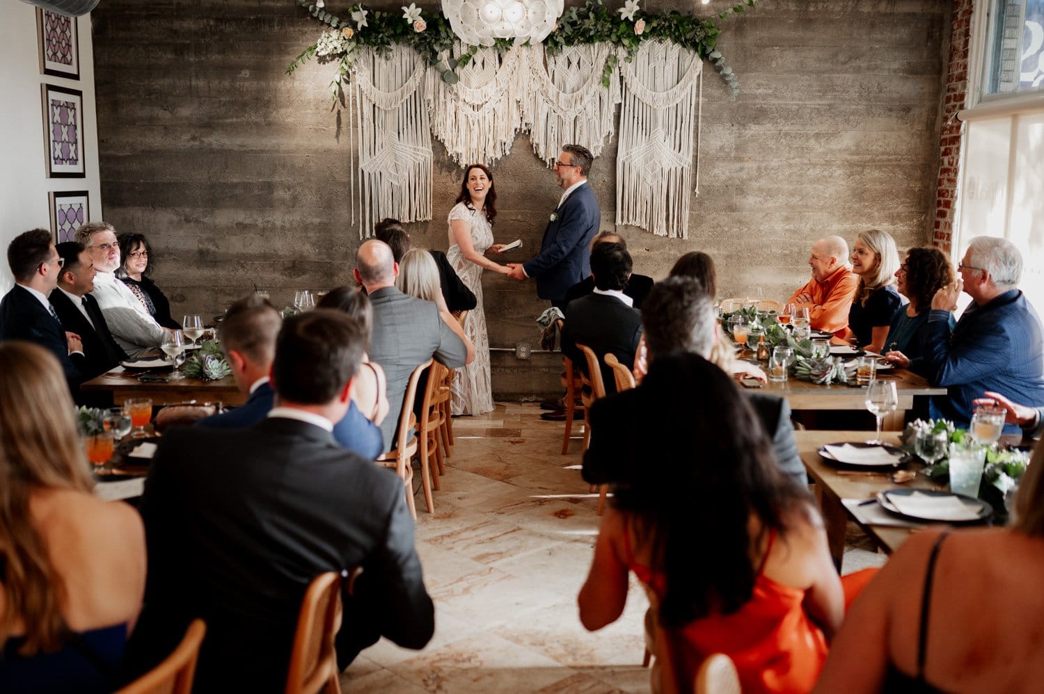 A couple exchanges vows at the boho-themed indoor wedding at Opal 28, with seated guests surrounding them. The decor features earthy tones, macrame hangings, and abundant greenery.