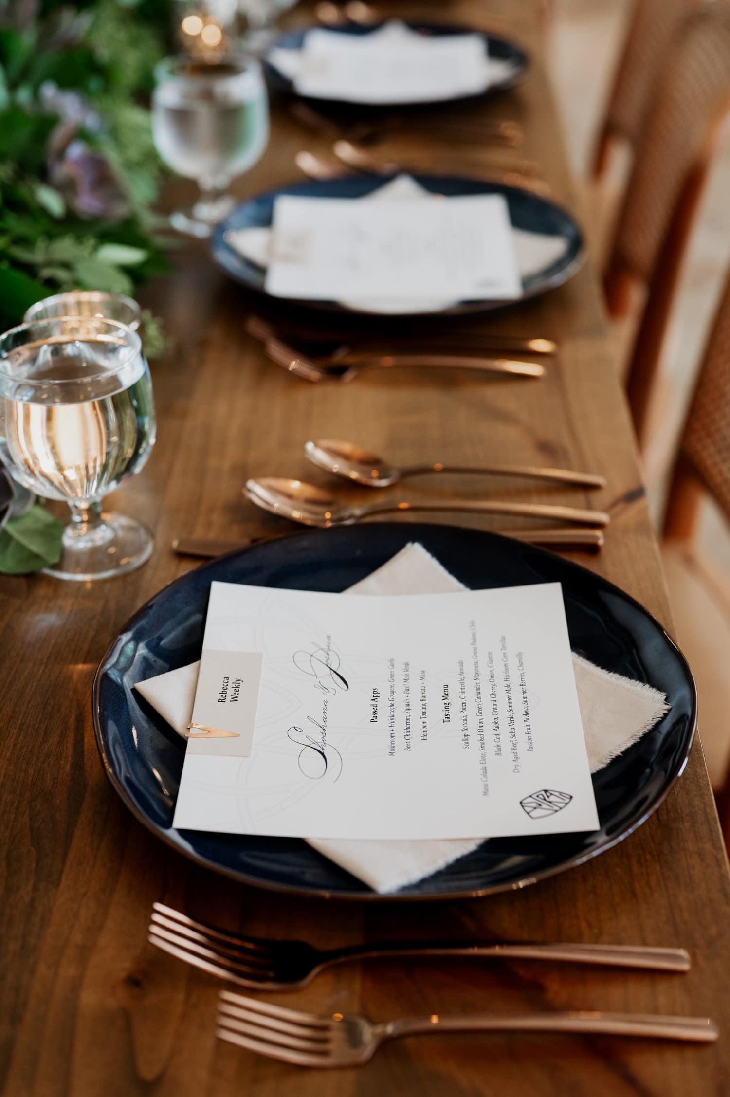 A beautifully arranged wedding table at Opal 28 with navy plates, menus, and gold cutlery set on a wooden table. Sparkling water glasses sit beside lush greenery for an elegant and refined look.