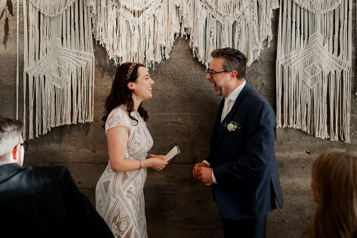 A bride and groom smile joyfully during their wedding ceremony at Opal 28, with a detailed macrame backdrop behind them.