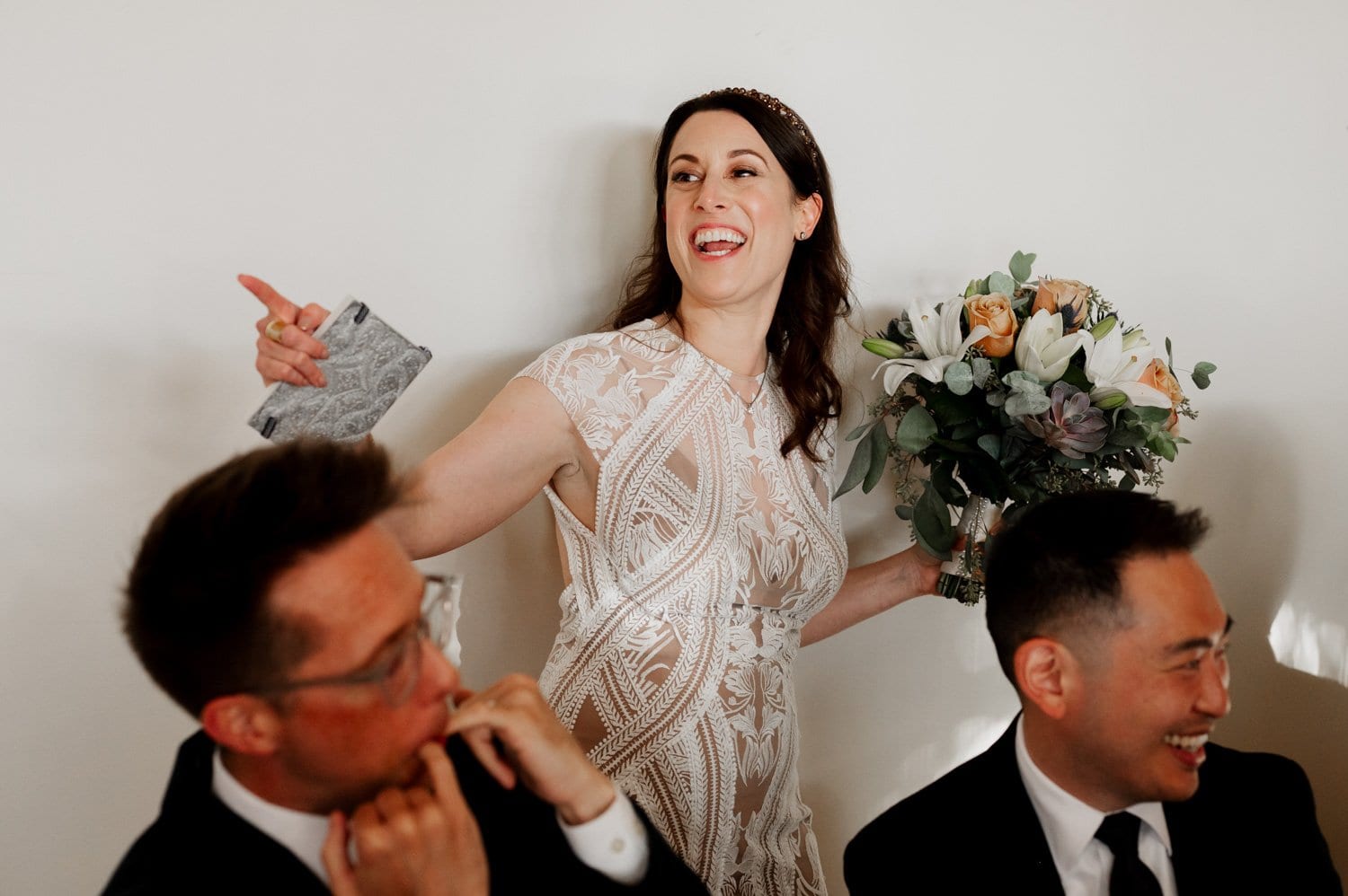 A joyful bride in a lace dress laughs with a bouquet in hand, gesturing animatedly while two seated men smile warmly beside her at a wedding at Opal 28.