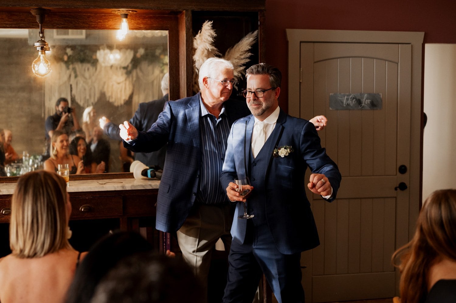 Two men in suits smile and laugh together at a wedding celebration, with other guests mingling in the background at Opal 28.