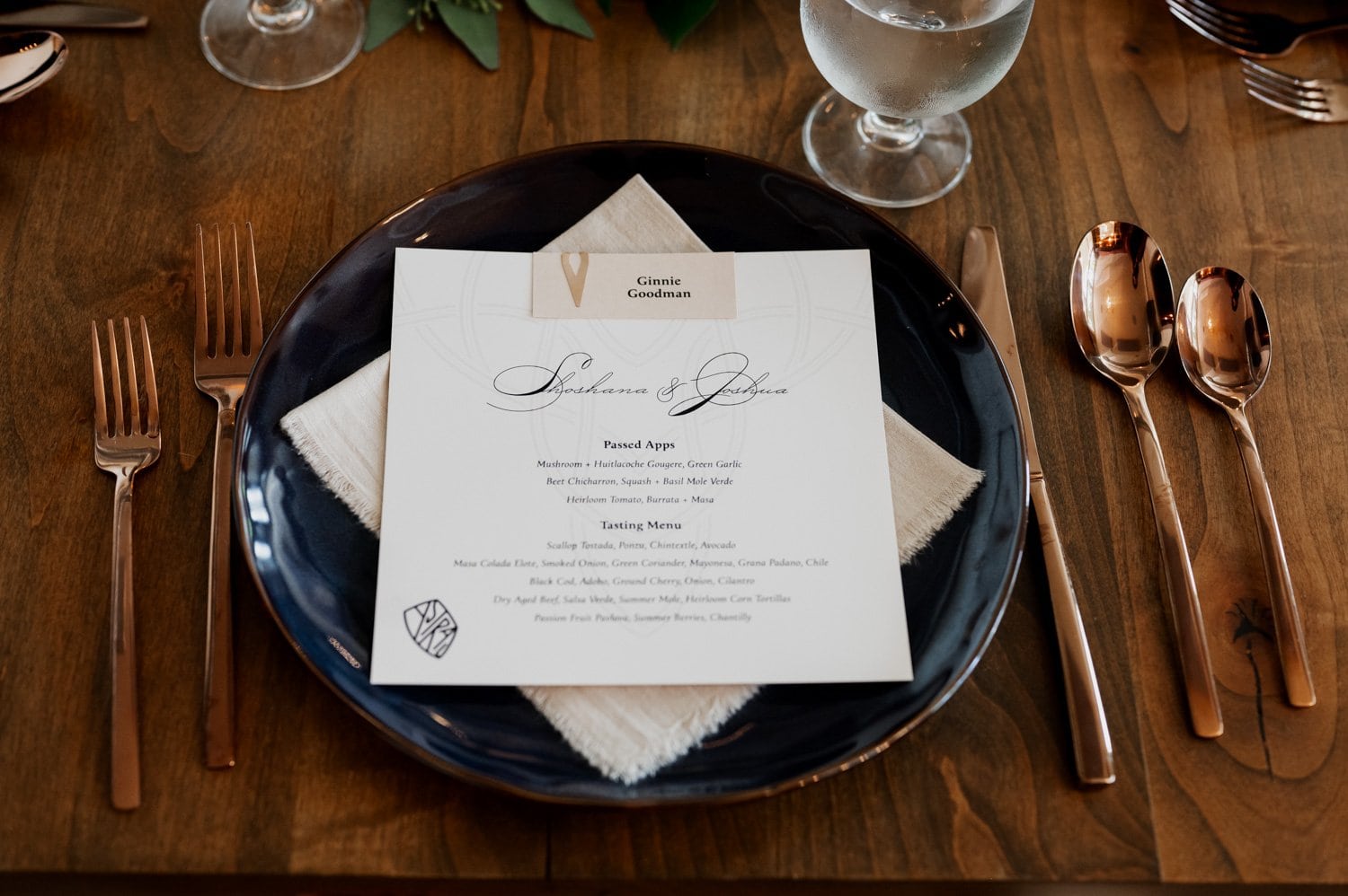 Elegant wedding table setting at Opal 28 with a menu card placed on a navy plate. The setup includes cutlery and a water glass arranged neatly on the wooden table.