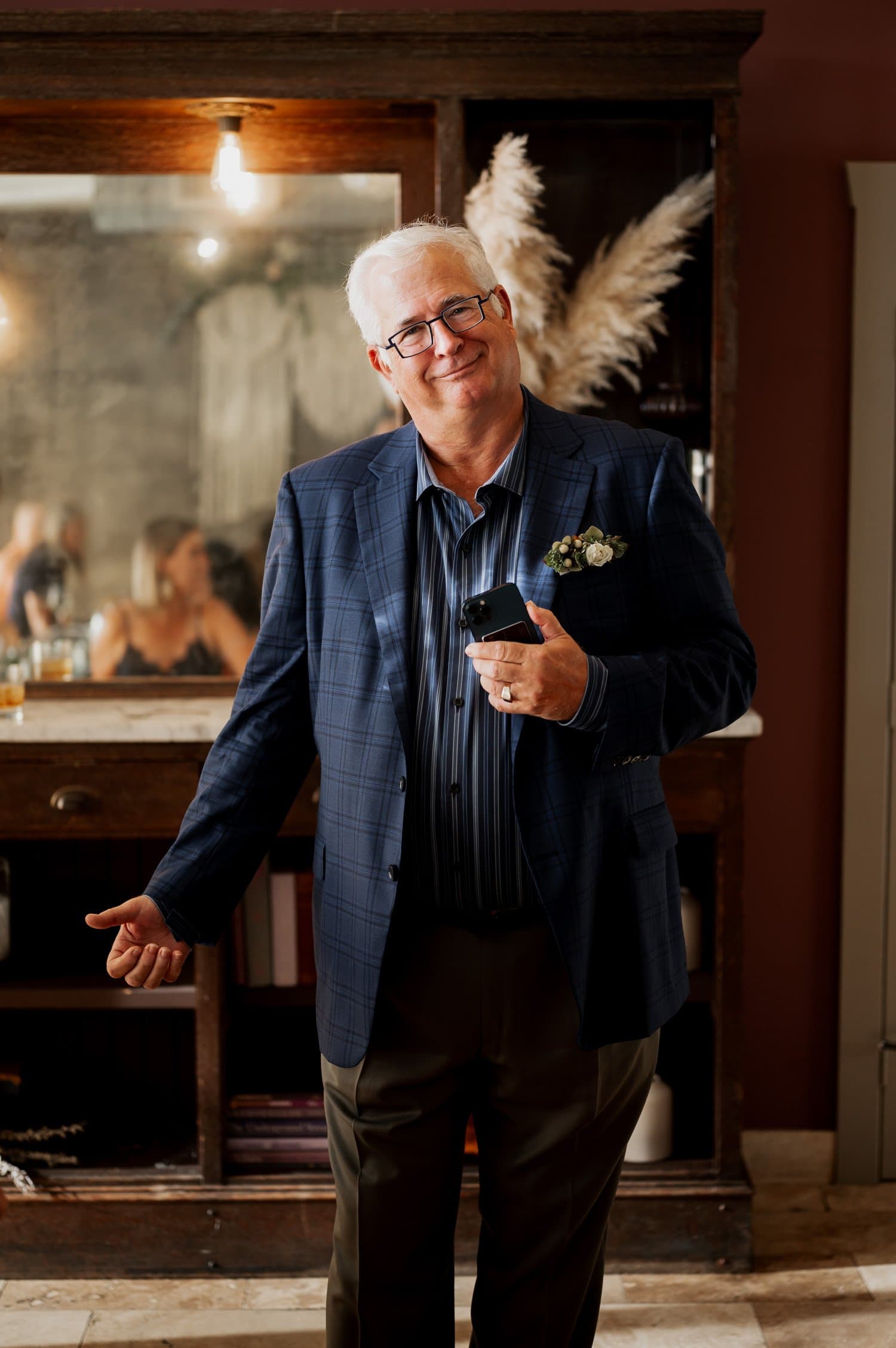 A man in a suit smiling while holding a phone, standing in a warmly lit room with pampas grass decorations in the background, possibly at an event or wedding hosted at Opal 28.