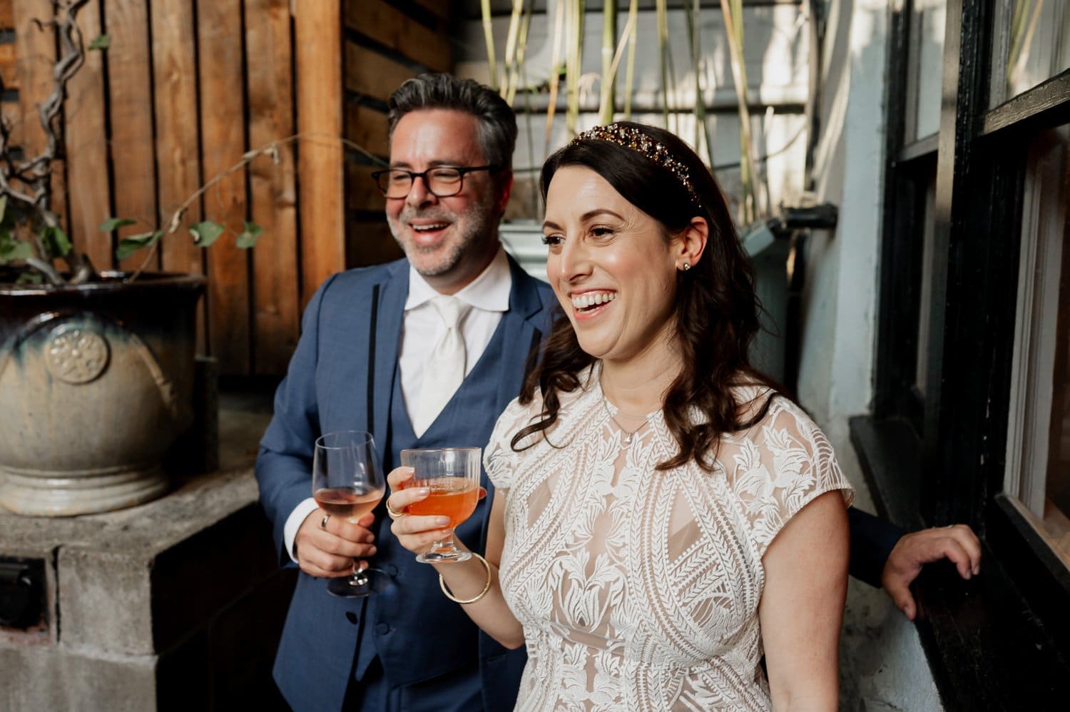 A joyful couple stands outdoors by a wooden wall and potted plant, holding drinks and smiling at their wedding at Opal 28.