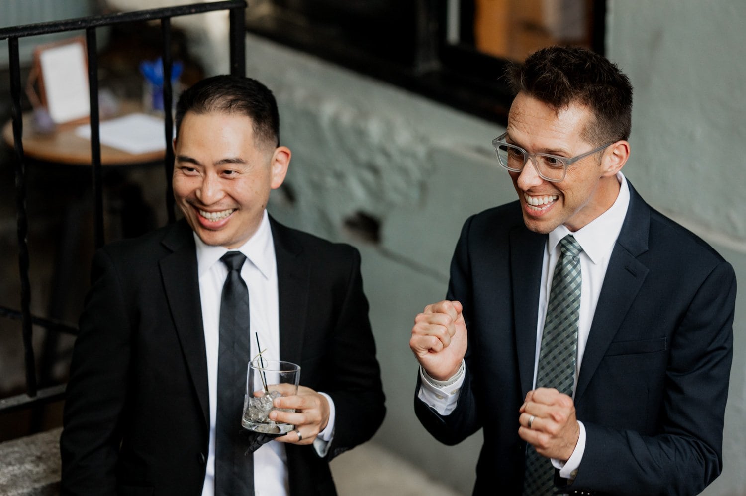 Two men in suits celebrating near a railing at Opal 28, with one man clenching his fists and the other holding a glass at a wedding.