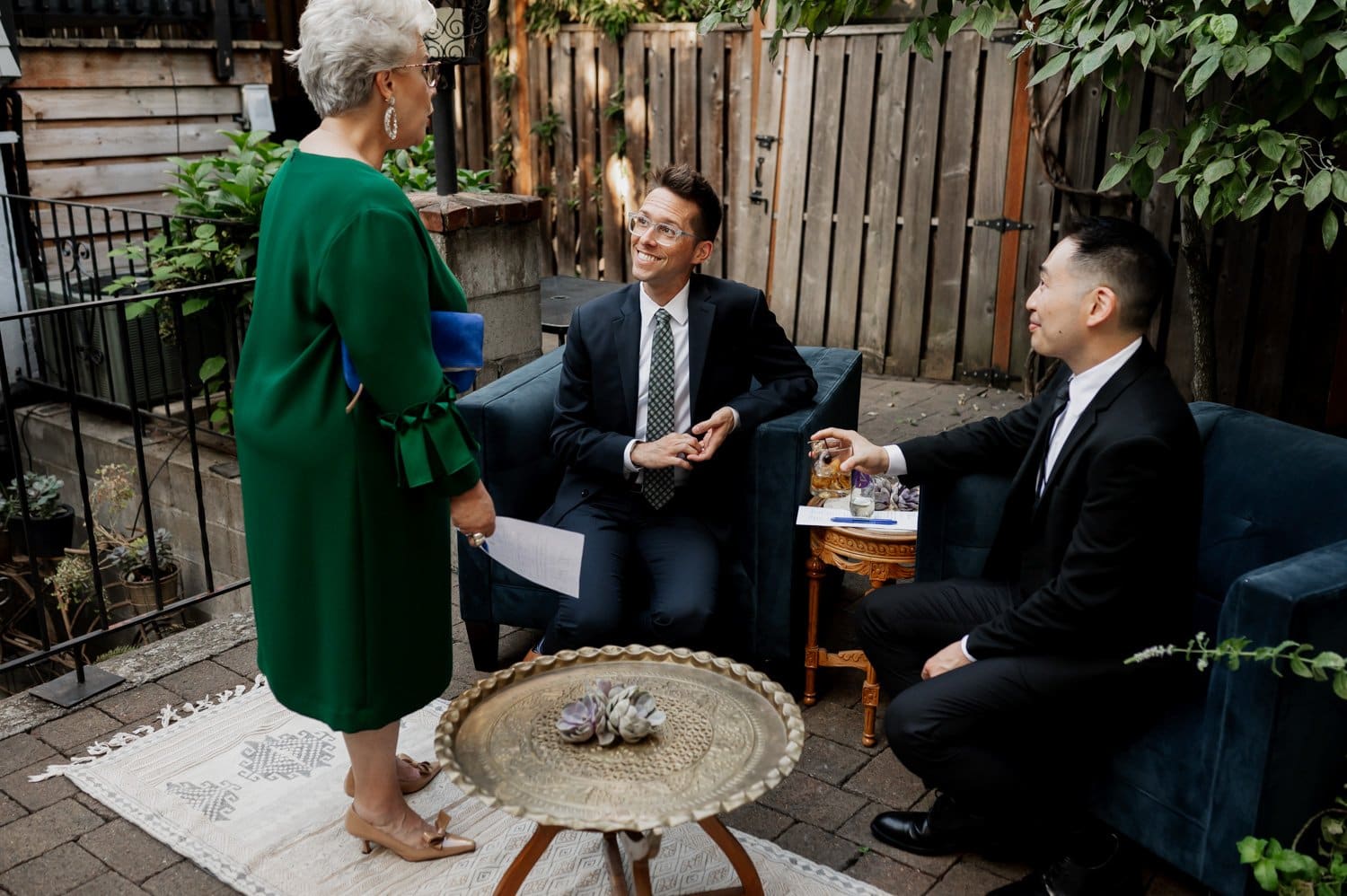 A woman in a green dress engages in conversation with two men wearing suits, seated outdoors at a wedding at Opal 28. A tray and vibrant plants enhance the foreground setting.