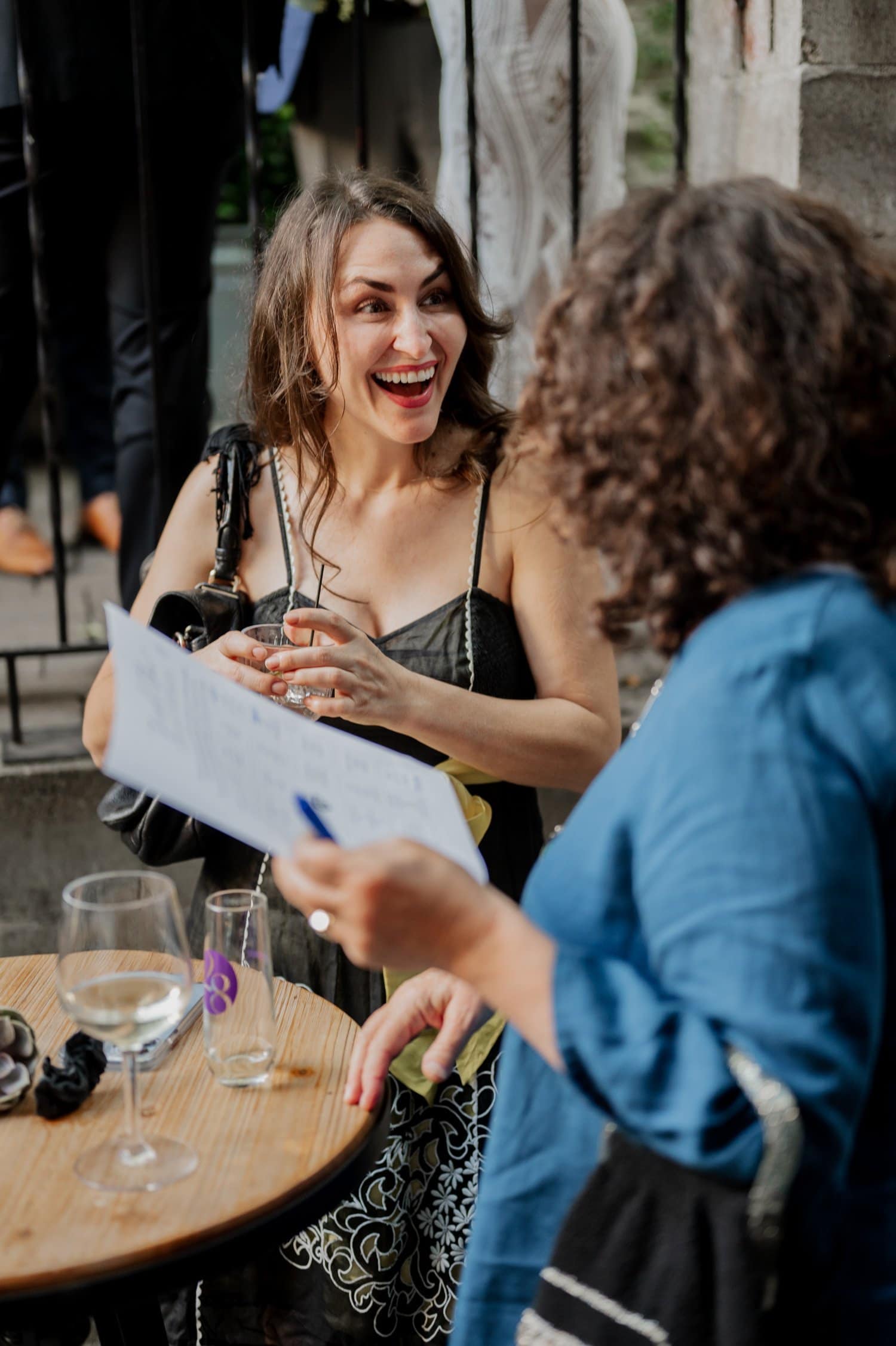 Two women are sitting at a table with drinks in an outdoor setting, engaging in animated conversation and laughter while discussing plans for a wedding at Opal 28.