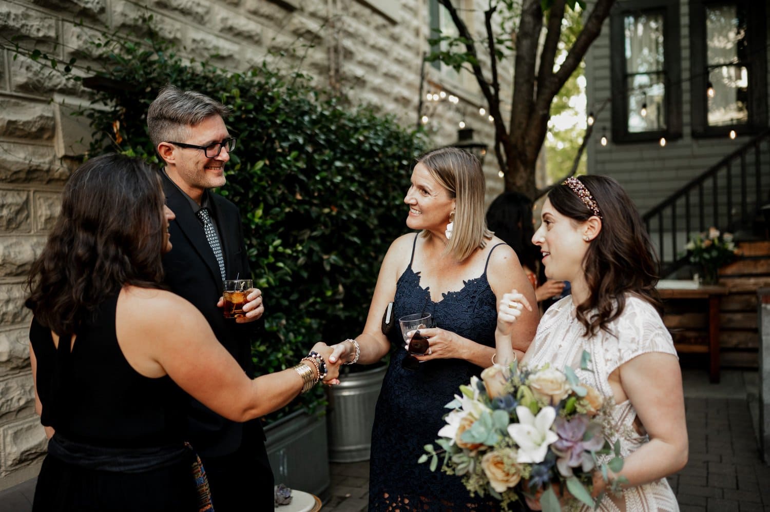 Four people in formal attire are chatting outdoors at a wedding at Opal 28. One person is holding a bouquet, and the others are sipping drinks. They are surrounded by lush greenery and elegant stone walls.