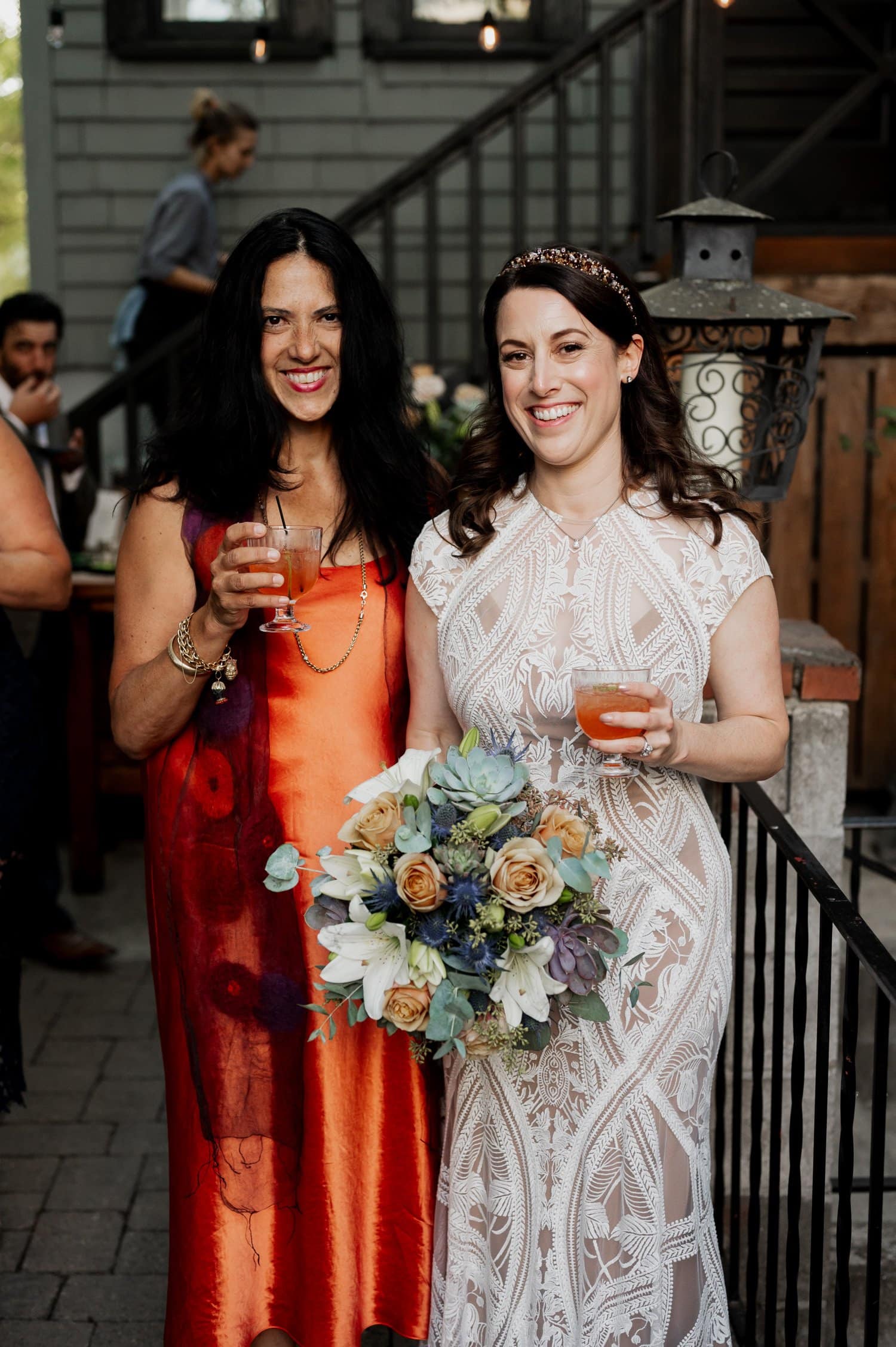 Two women smiling and holding drinks at a wedding; one in a patterned red dress, the other in a white lace dress with a bouquet, at Opal 28.