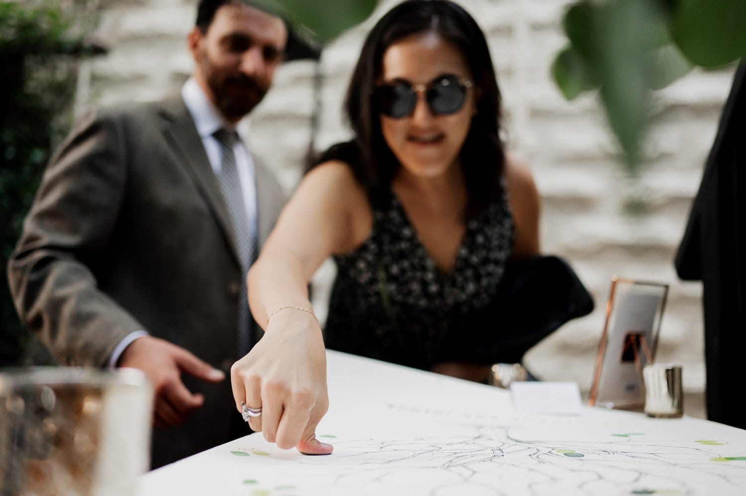A woman wearing sunglasses points at a seating chart while a man in a suit stands beside her, poised to help. They appear to be organizing an event at Opal 28.