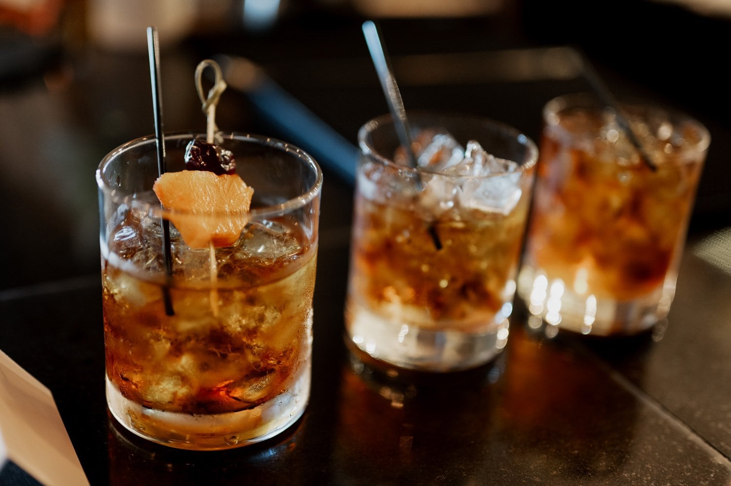 Three cocktail glasses on a bar, each garnished with cherries and orange twists, under soft lighting.