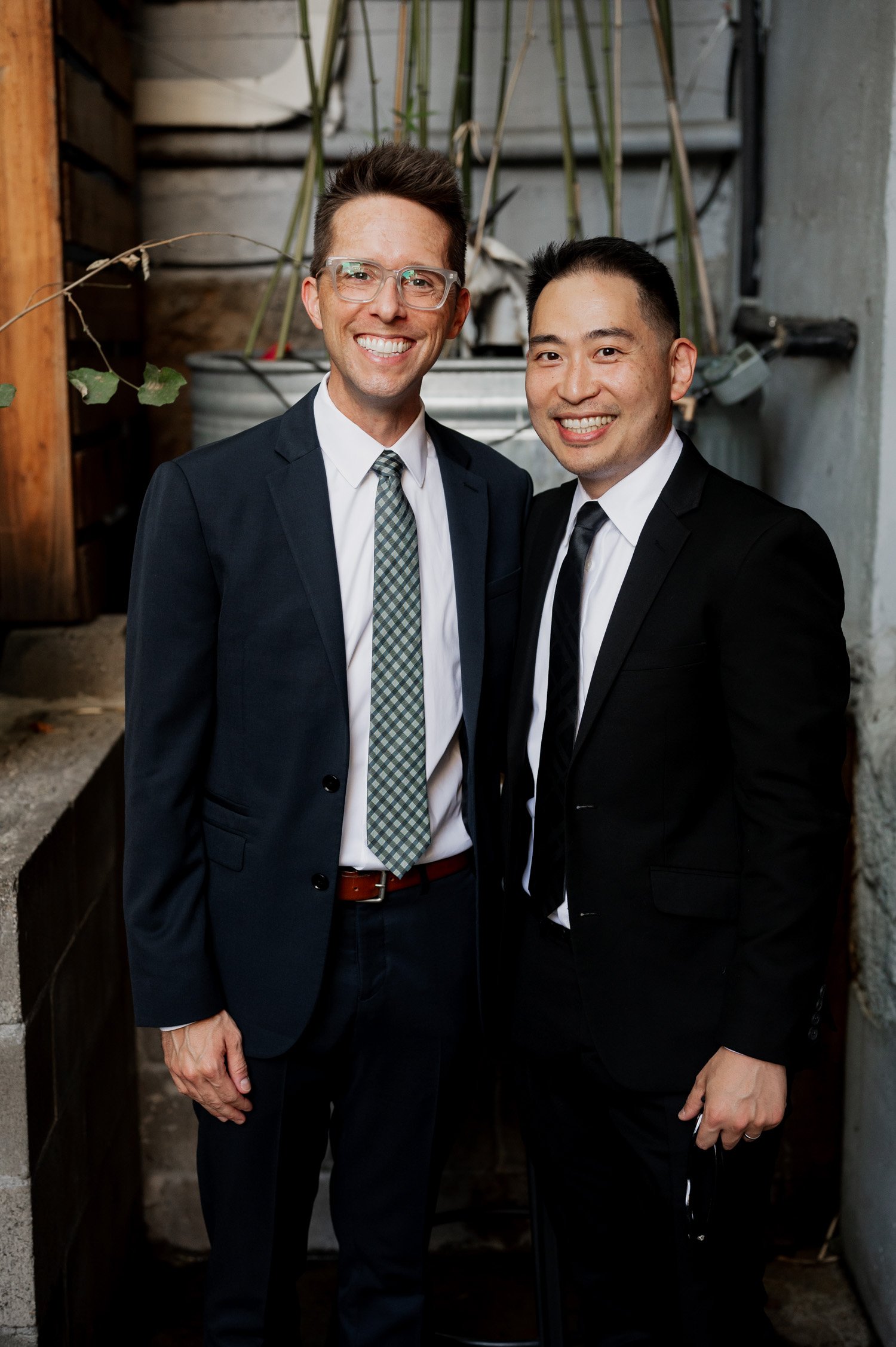 Two individuals in suits smiling and standing closely together, celebrating a special occasion. They are surrounded by vibrant urban elements and lush plants, indicating a lively atmosphere similar to a wedding at Opal 28.