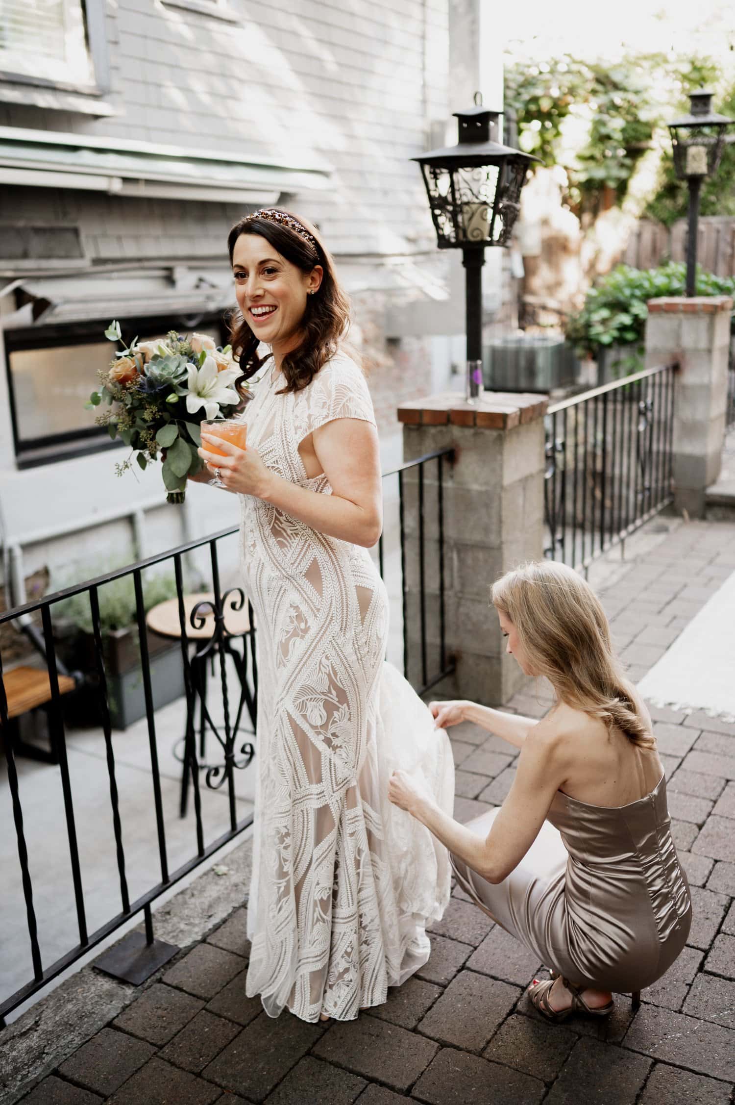 A bride smiles while holding a bouquet and a drink as a woman adjusts her dress on a brick patio surrounded by greenery at Opal 28 during their wedding.