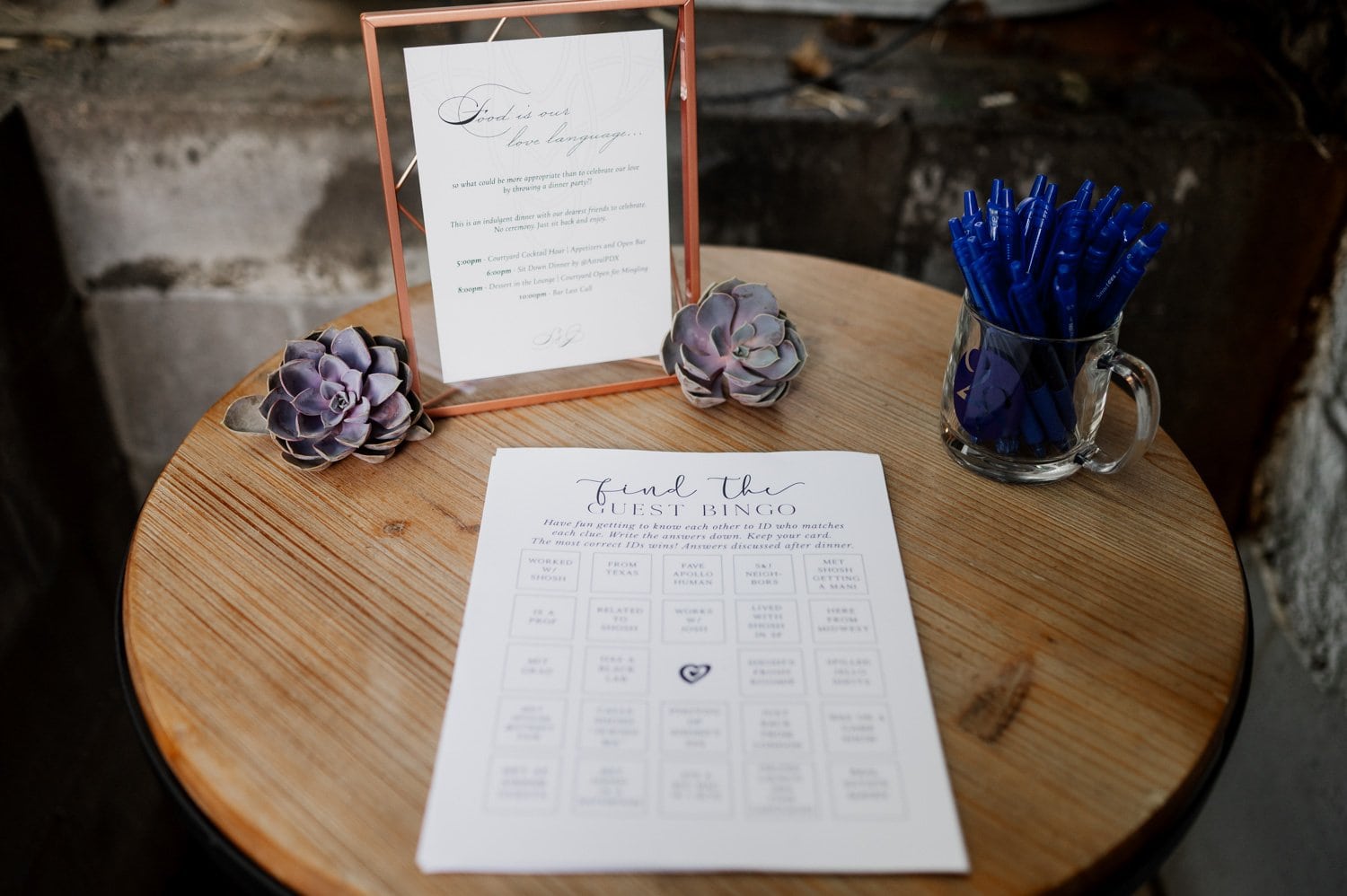 A bingo card sits on a wooden table next to a mug filled with blue pens and a framed sign, surrounded by purple succulents at the wedding venue Opal 28.