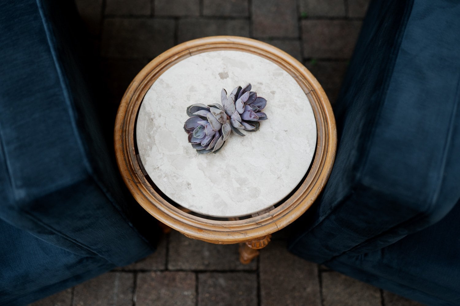 A charming round wooden table with succulents on top, surrounded by two dark blue chairs, placed on a brick patio, creating a wedding-like setting at Opal 28.