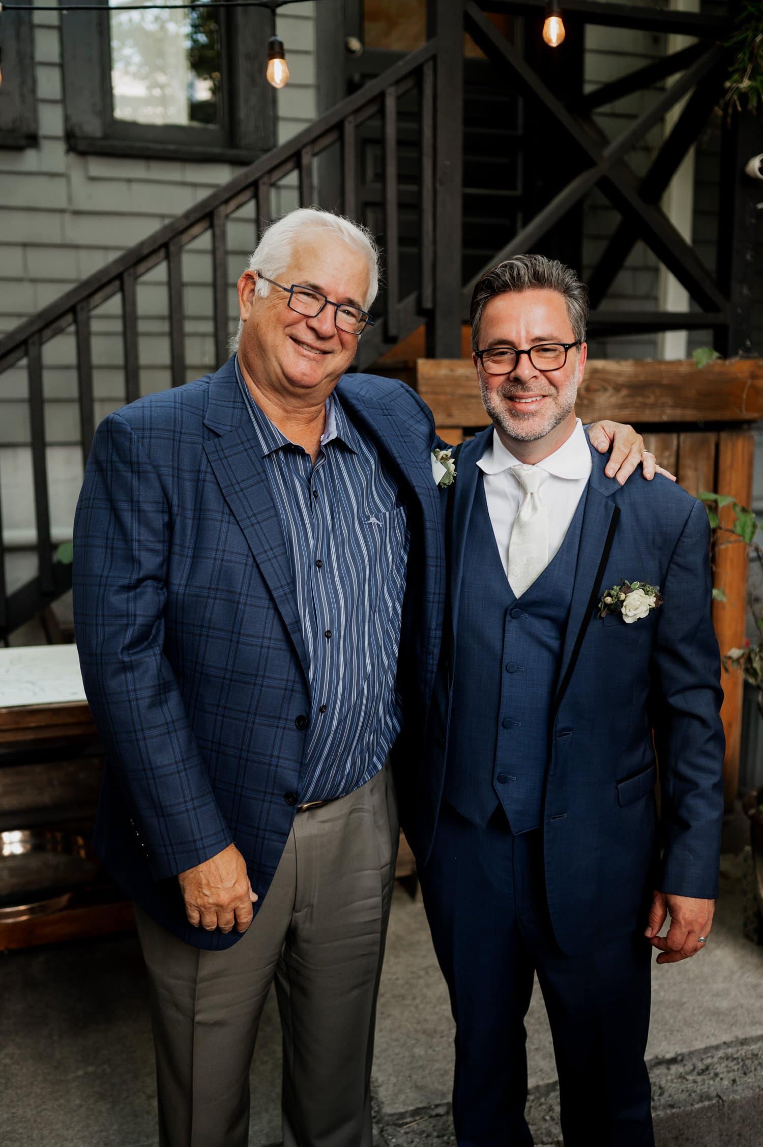 Two men in suits standing together and smiling in an outdoor area with a wooden backdrop.