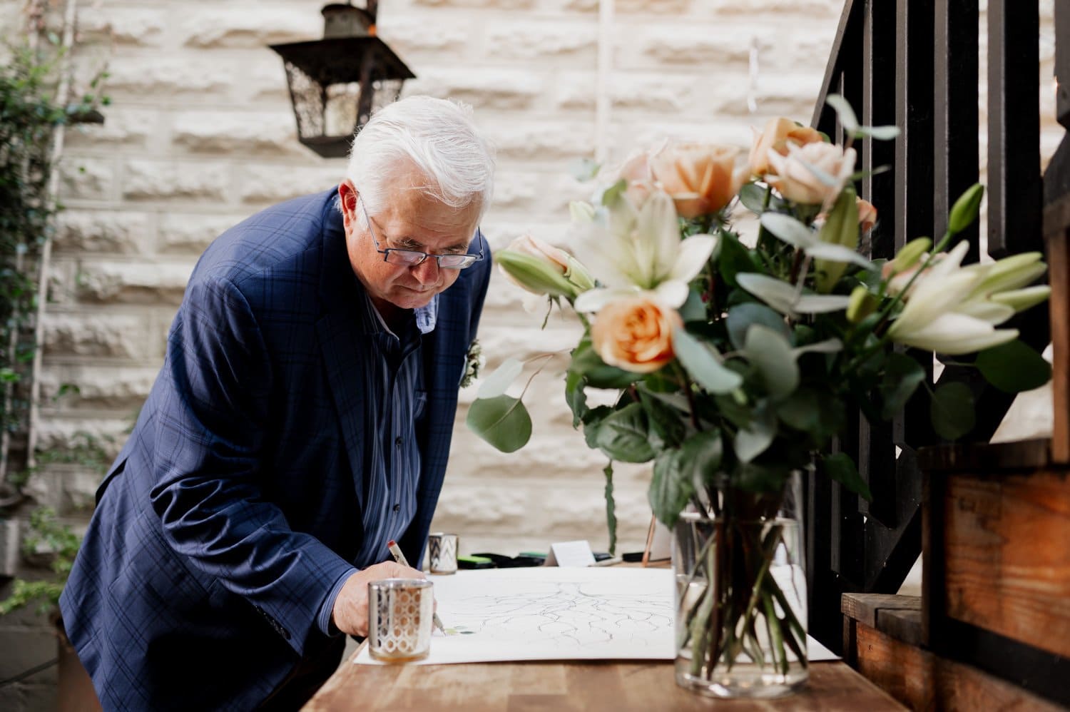An elderly man in a blue suit signs a book at a wooden table with flowers, part of a wedding setting at Opal 28.
