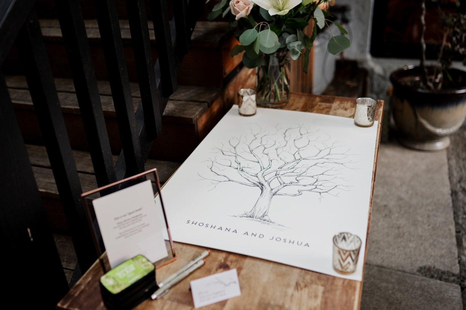 Poster featuring a bare tree illustration with Shoshana and Joshua text, designed for a wedding at Opal 28. The artwork is displayed on an elegantly set table adorned with flowers and candles.