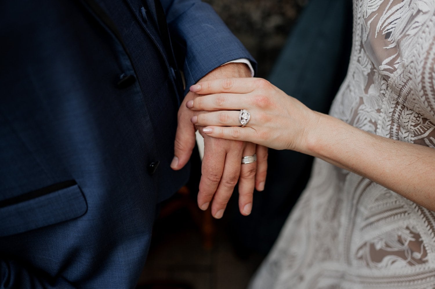 Close-up of a couple holding hands, displaying their wedding rings. The woman is wearing a lace dress, and the man is in a blue suit. They are at Opal 28.