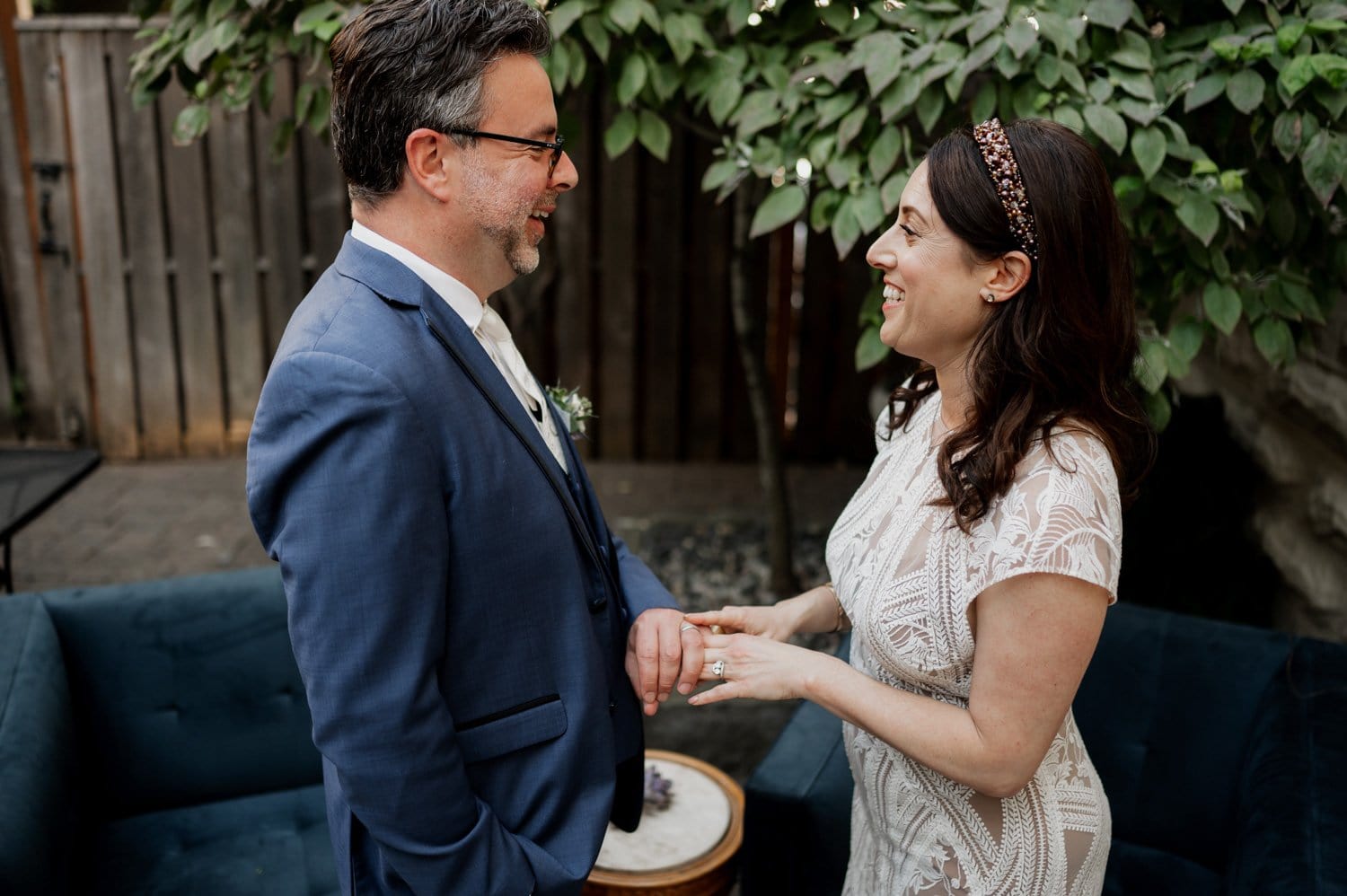 A couple smiles at each other while holding hands in a leafy outdoor setting, with a blue couch and wooden fence nearby.