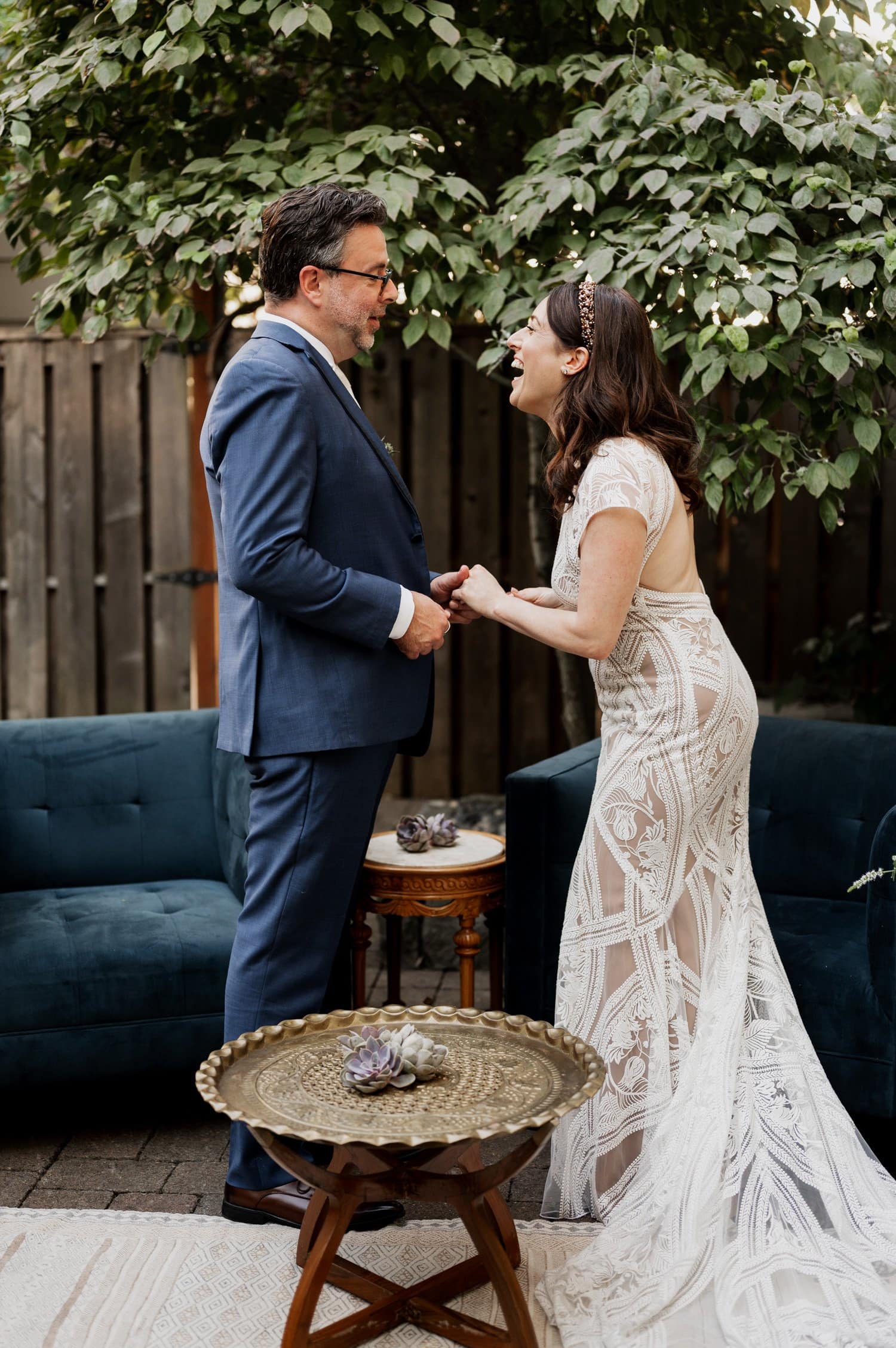 A couple smiles and holds hands during their wedding at Opal 28, surrounded by lush greenery and dark blue decor.