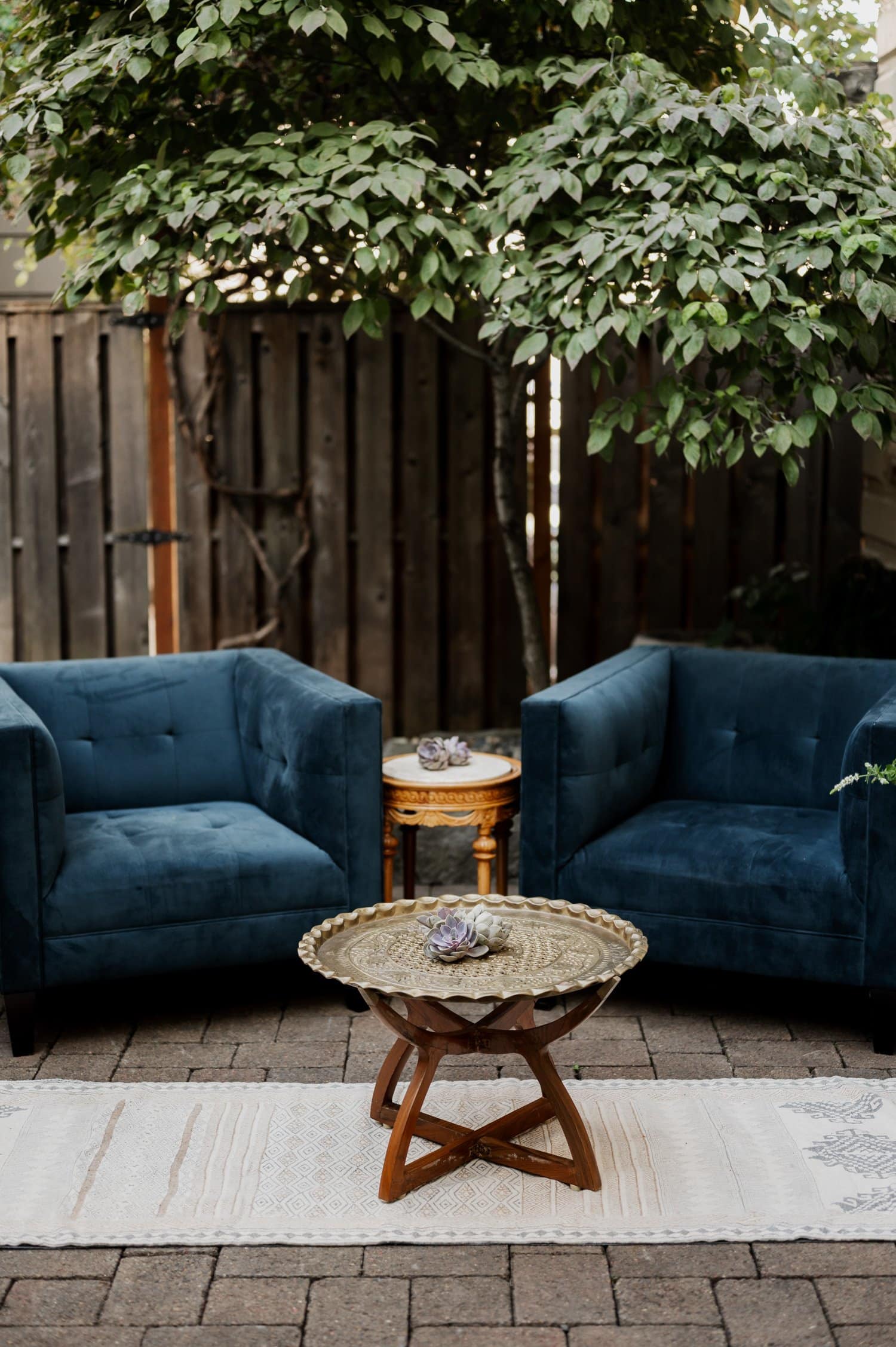 Two blue armchairs and a decorative table on a patio rug, set against a wooden fence and leafy tree, create an intimate setting for conversations during a wedding at Opal 28.