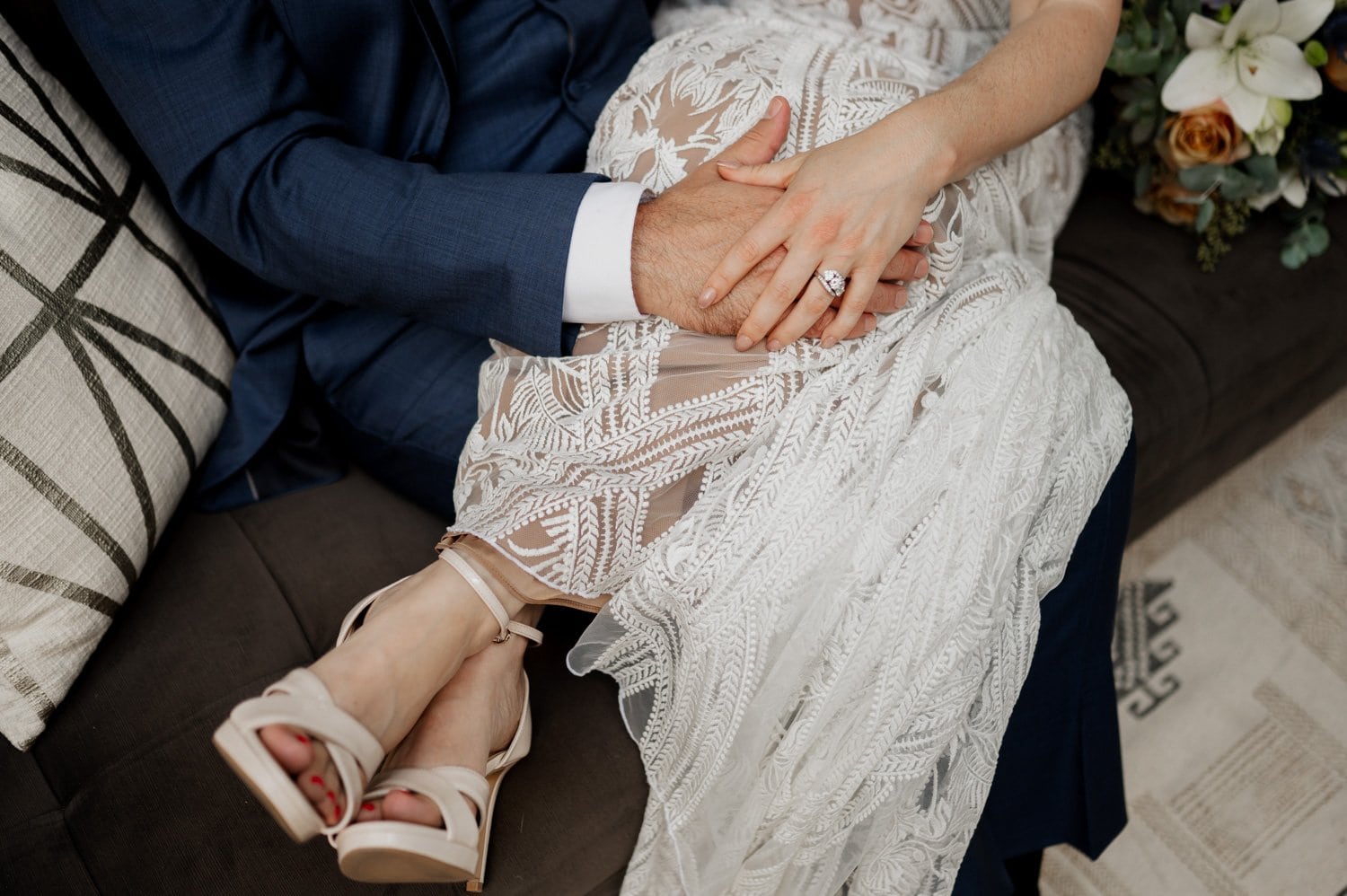 A couple sits on a couch holding hands, with her wearing a lace dress and heels; a bouquet rests beside them, capturing a peaceful moment from their wedding at Opal 28.