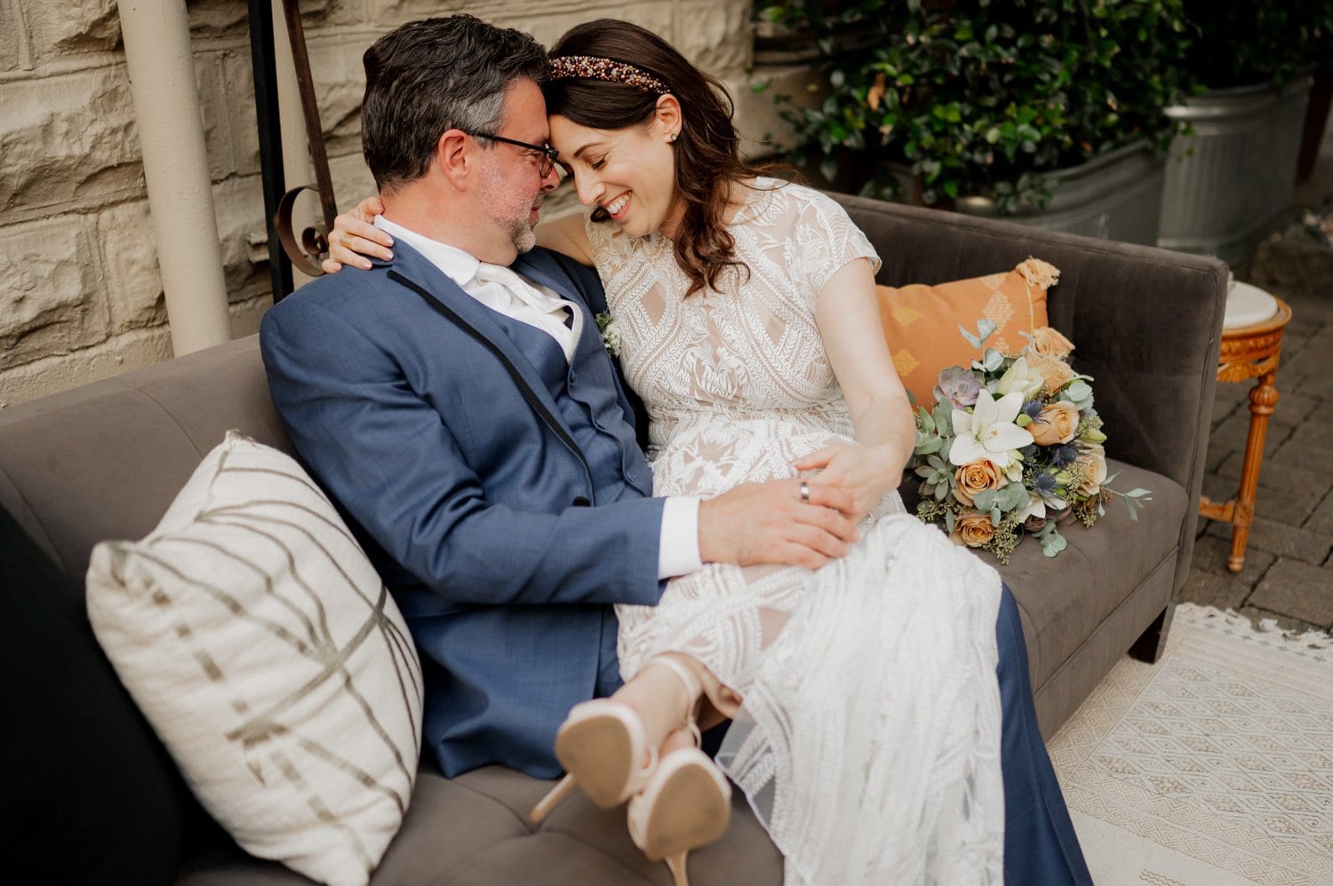 A couple in wedding attire sits closely on a sofa at Opal 28, smiling affectionately with a bouquet resting nearby.