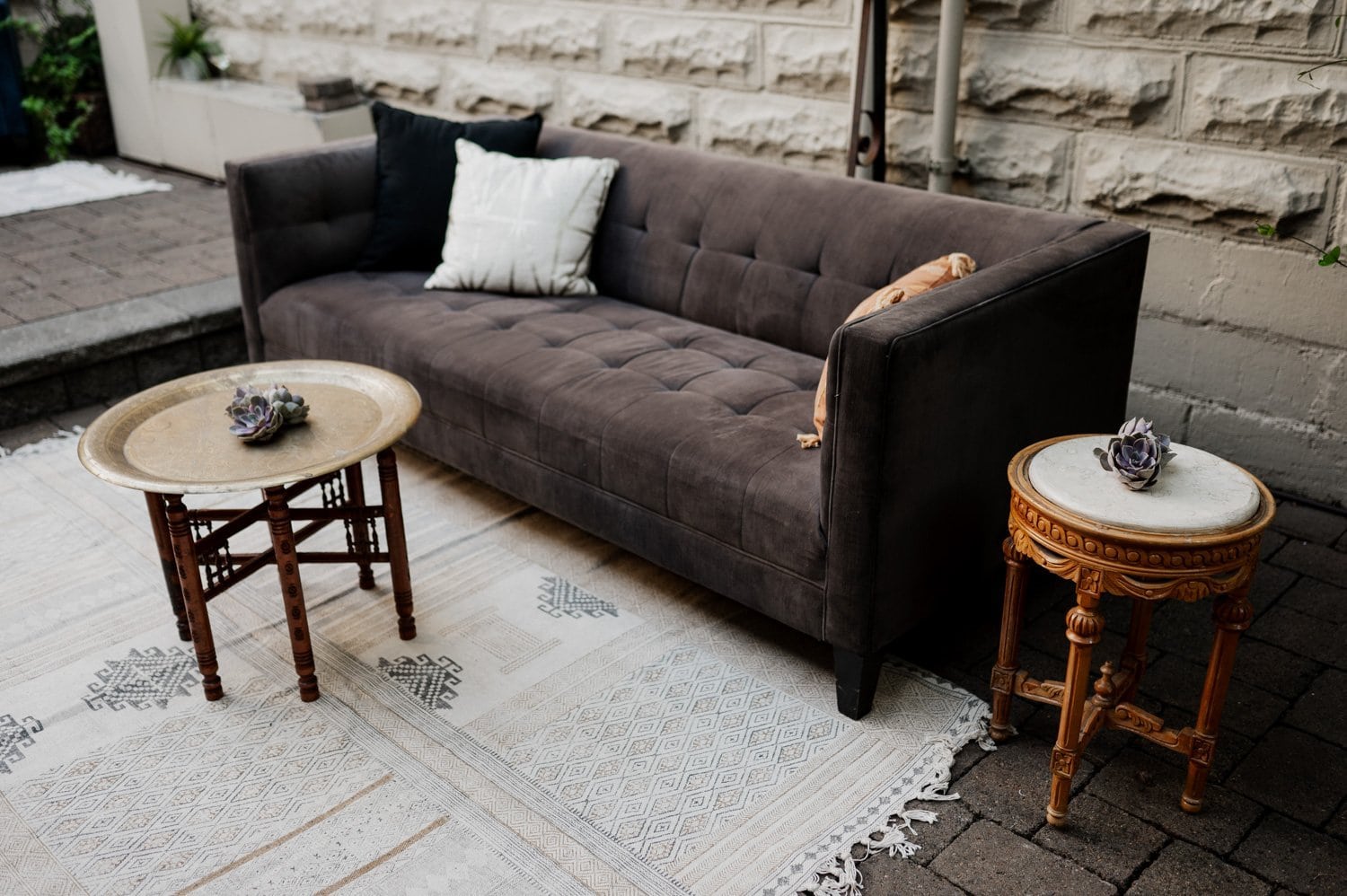 Outdoor seating area at Opal 28, with a gray couch, two small round tables, and decorative pillows on a patterned rug. Ideal setting for a wedding event.