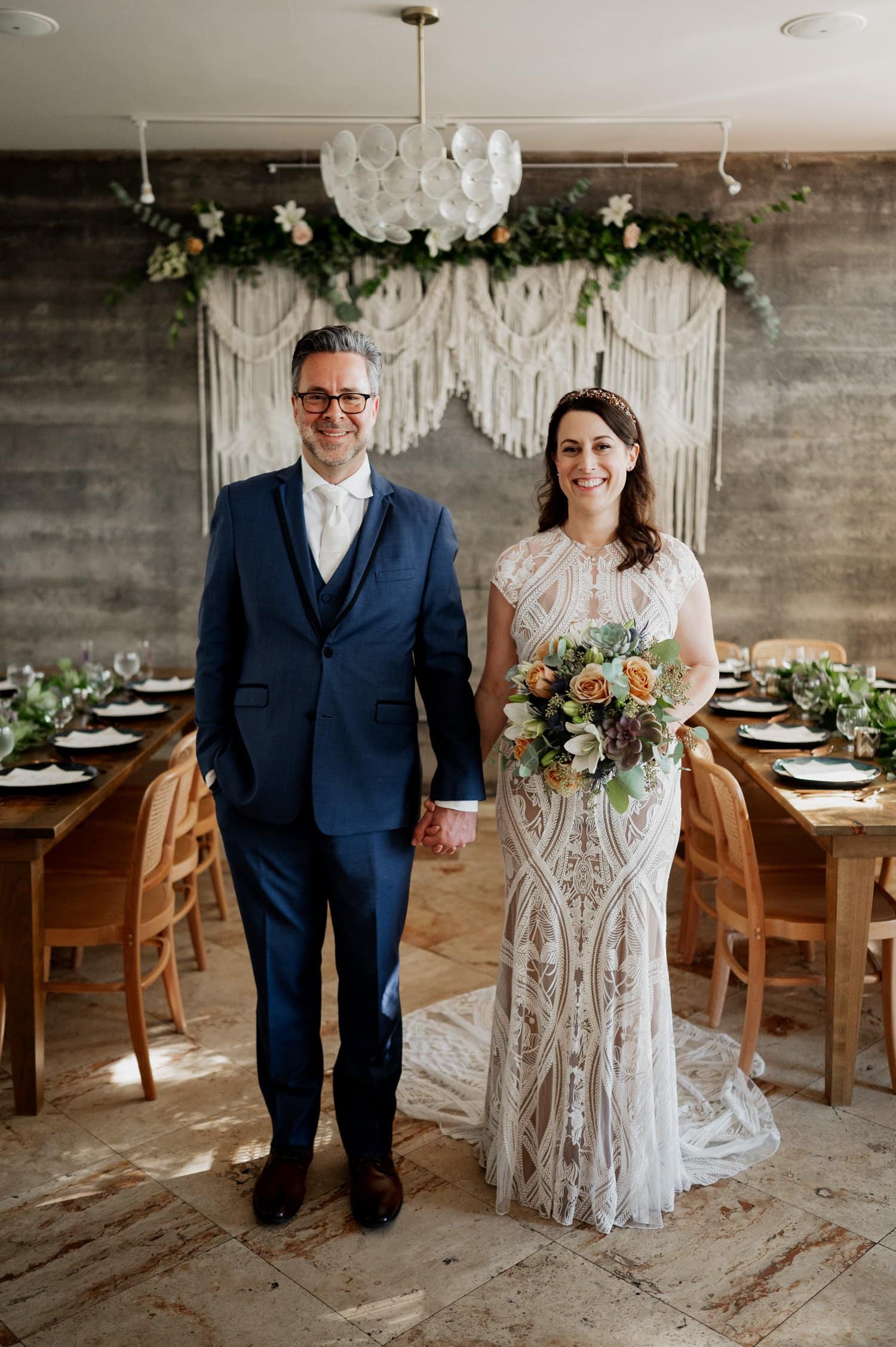 The couple, dressed in elegant wedding attire, stands before a beautifully adorned table of lush greenery and flowers at their enchanting wedding at Opal 28, holding a stunning bouquet.